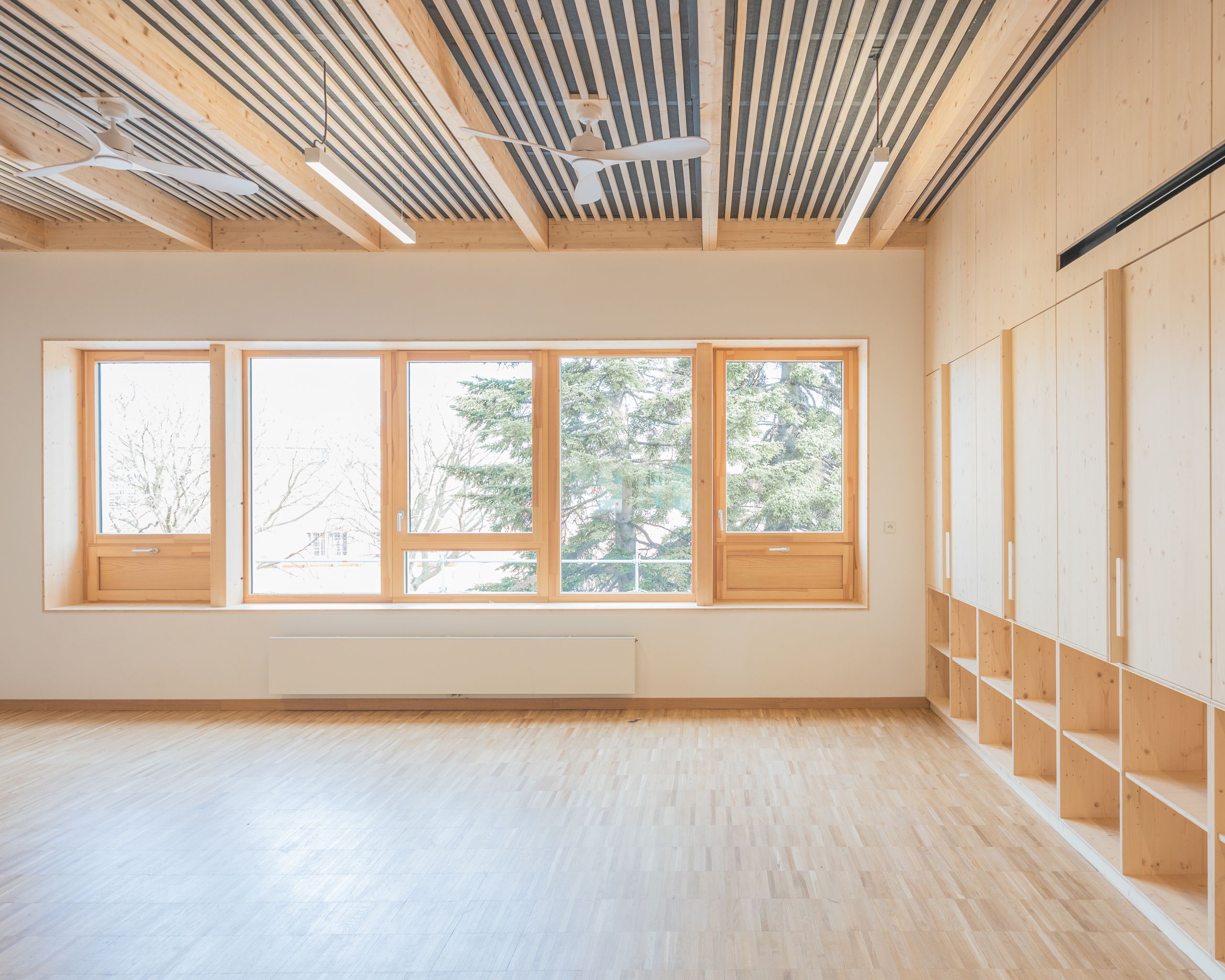 Intérieur du Groupe scolaire J.F. Kennedy à Lyon : salle lumineuse en structure bois apparente, plafond technique, grands châssis vitrés et rangements intégrés.