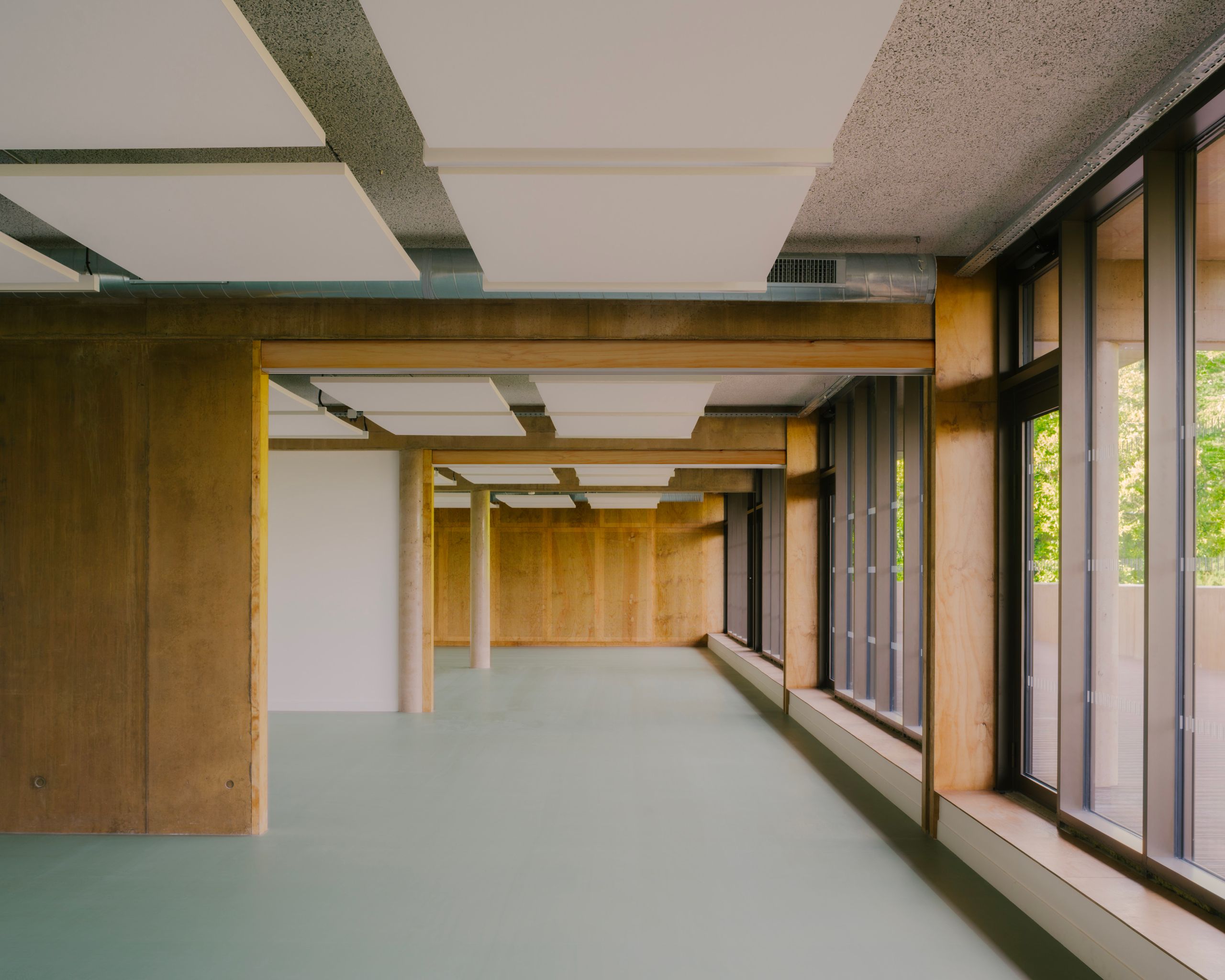 Intérieur de la Maison des familles à Limonest : plateau libre en béton et bois, grandes baies vitrées sur jardin, plafond à dalles acoustiques suspendues.