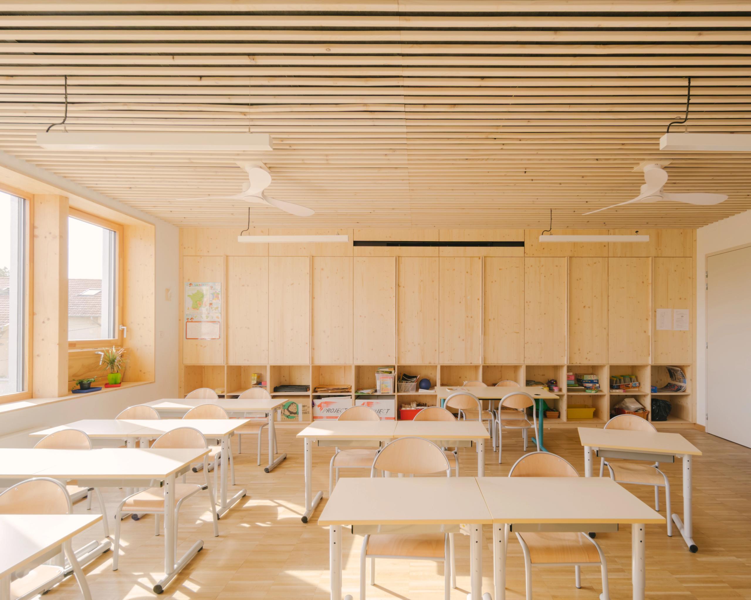 Salle de classe du groupe scolaire Kennedy à Lyon, structure bois apparente, plafond à lames, grands châssis latéraux et mobilier clair modulable.