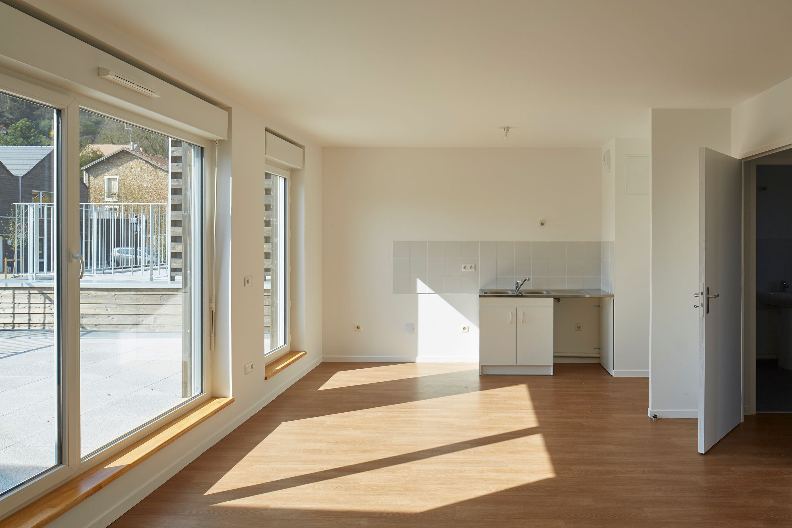 Intérieur d’un logement des 36 logements à Vauhallan par Tectoniques, pièce principale traversante avec grandes baies vitrées, sol bois et kitchenette intégrée.