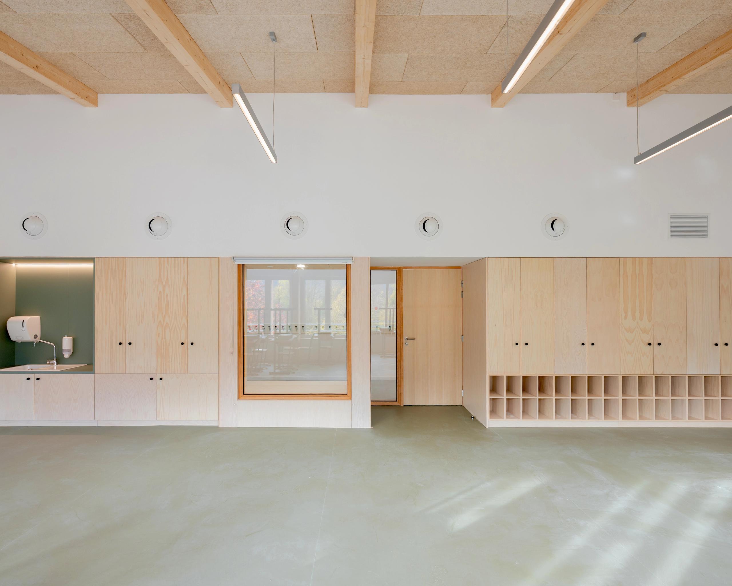 Intérieur du groupe scolaire du Bouchet à Bonneville : salle claire avec mobilier en bois, rangements intégrés, menuiseries vitrées et plafond à poutres apparentes.