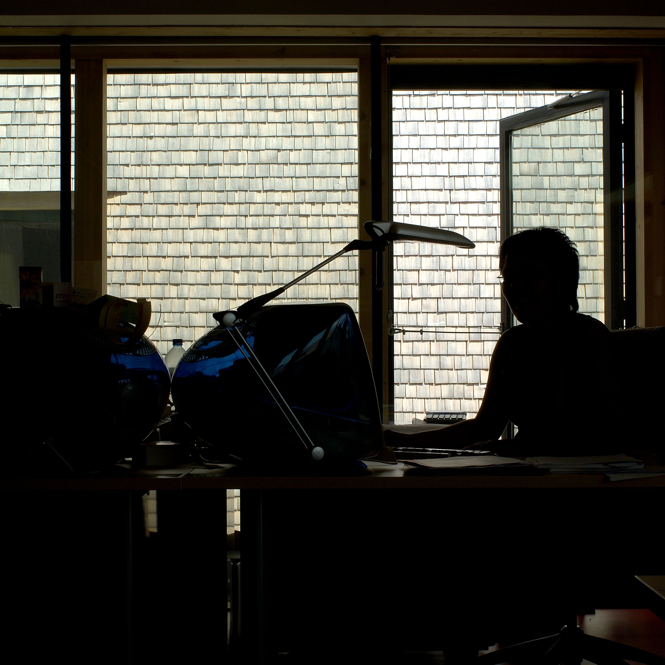 Intérieur de la Maison du Parc du Haut-Jura à Lajoux, bureau en contre-jour face à la façade en tavaillons d’épicéa et à une porte-fenêtre ouverte.