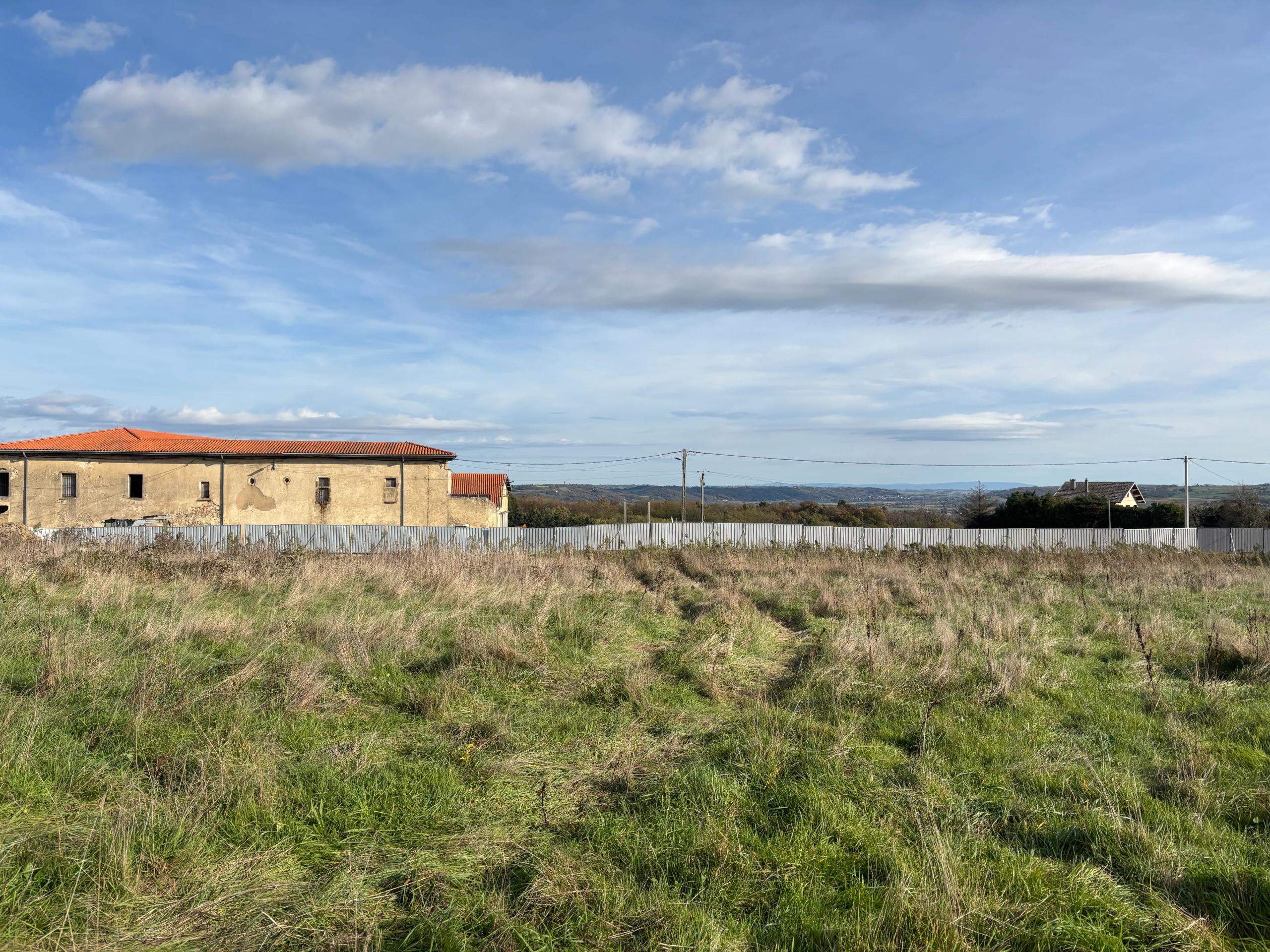 Vue du site du futur school complex à Vienne : prairie en pente, bâtiment existant en maçonnerie enduite et tuiles, horizon dégagé sur le paysage lointain.