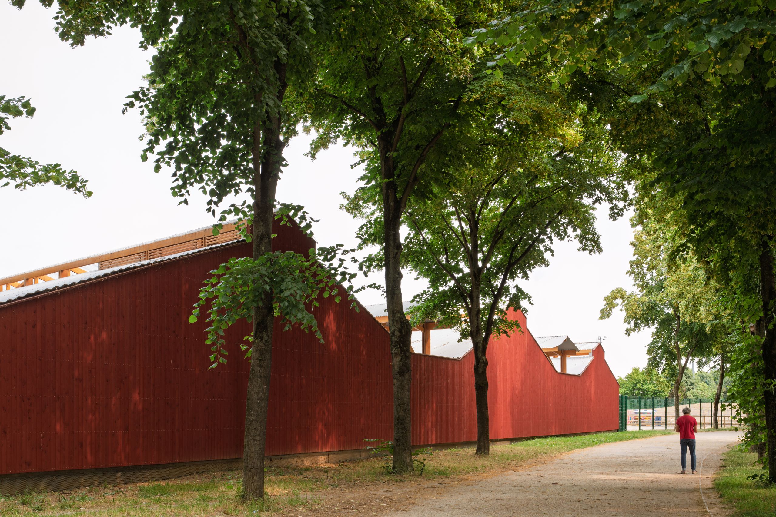 Centre équestre de Gennevilliers : long volume en bois bardé de rouge de Falun, toitures en sheds, aligné le long d’une allée plantée en lisière du parc.
