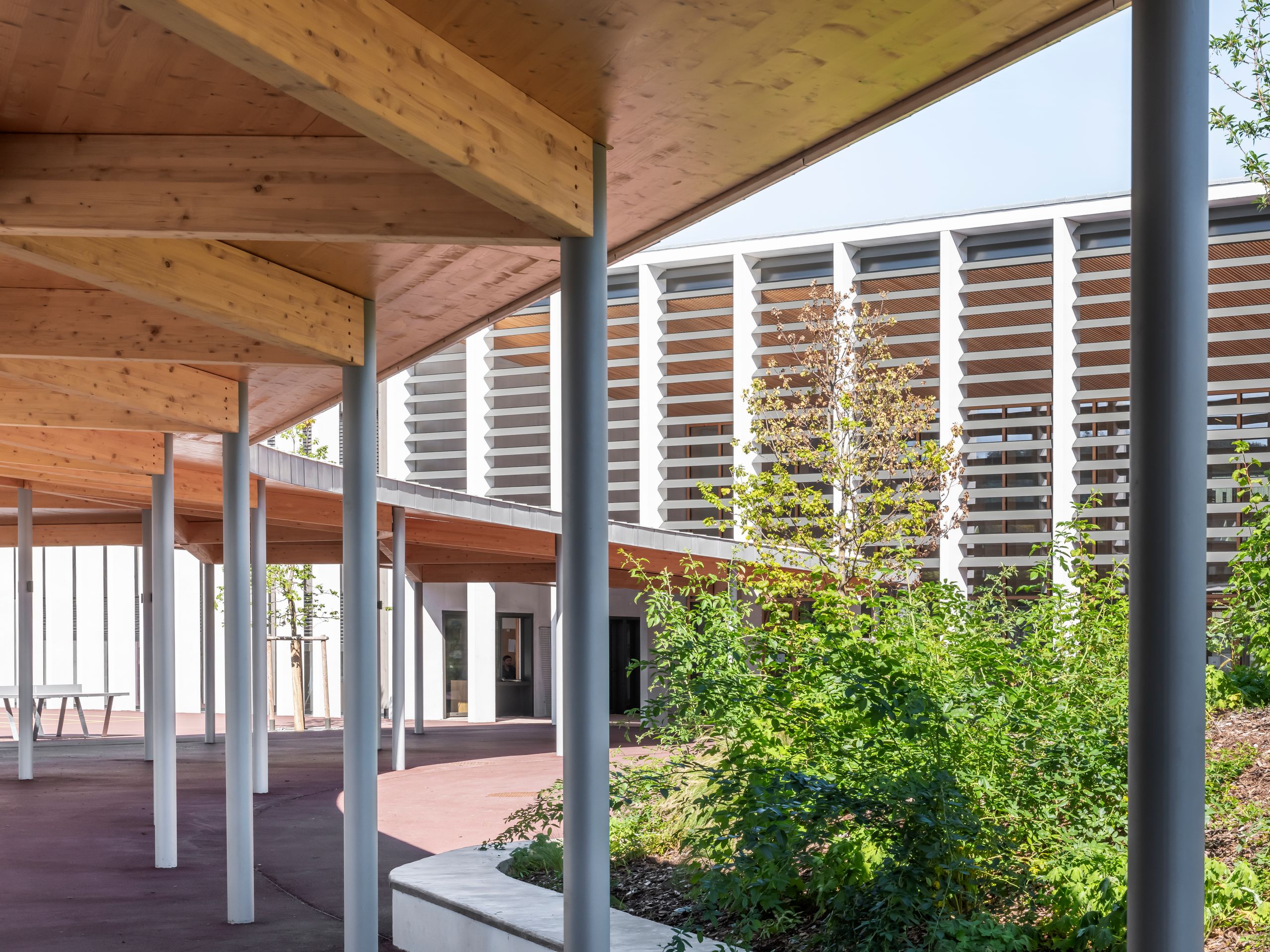 Cour du Collège du Ban à Vagney : préau en structure bois sur poteaux métalliques, façade à brise-soleil horizontaux et jardin planté en premier plan.