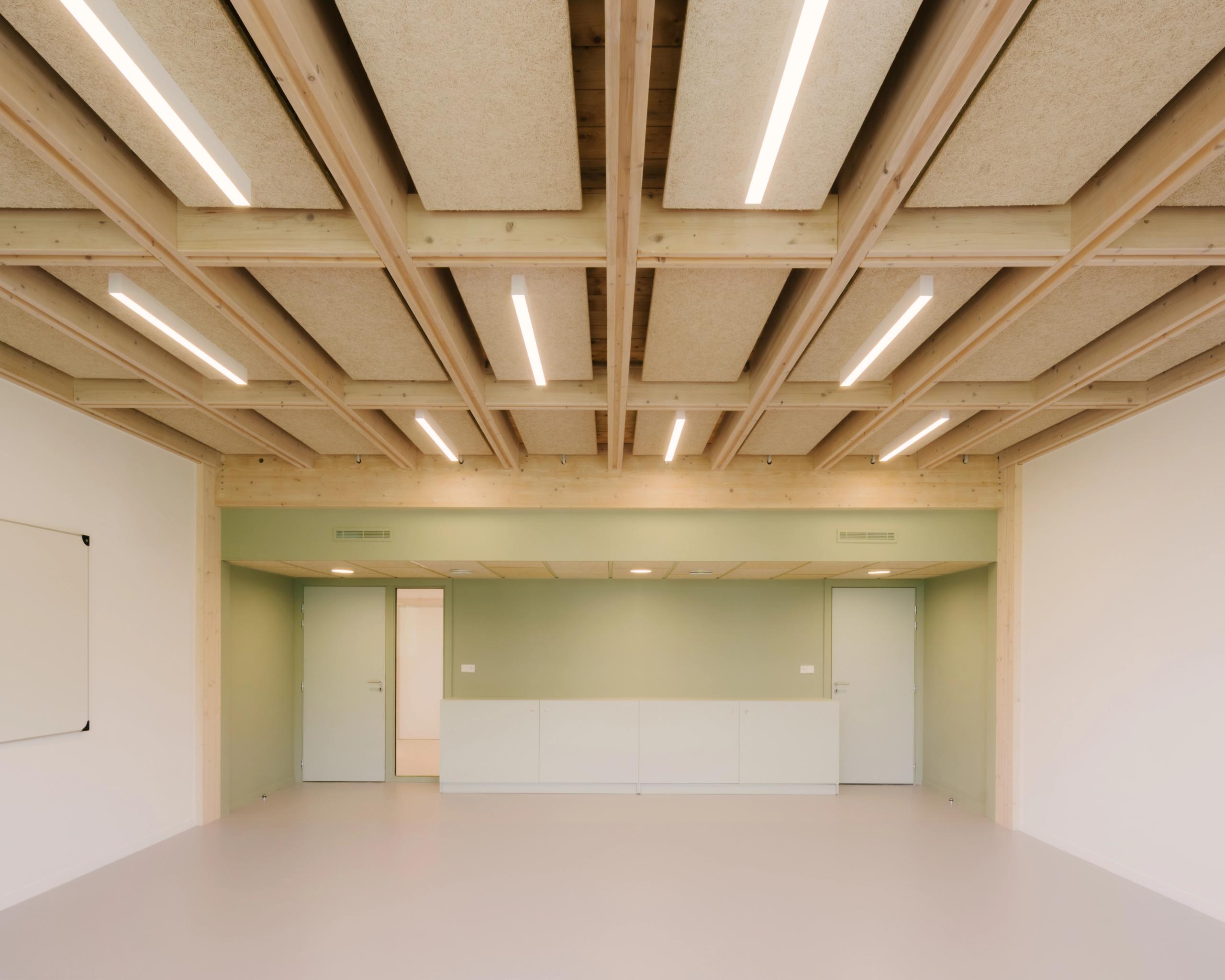 Salle du collège Maryse Bastié à Dole, restructurée par Tectoniques : plafond en caissons bois apparents, éclairage linéaire, parois vert pâle et sol clair.
