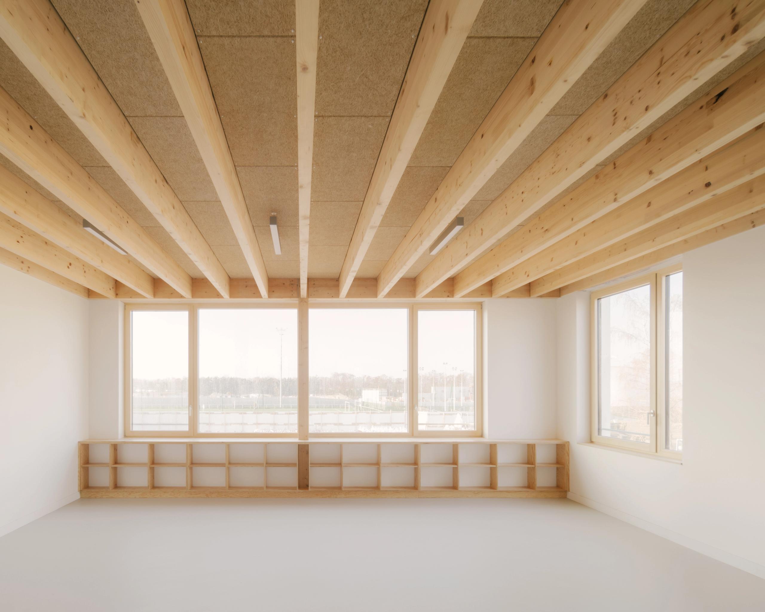 Salle des groupes scolaires Jacqueline Auriol et Jean Jaurès au Bourget, avec plafond à poutres bois apparentes, grandes baies vitrées et rangements bas intégrés.