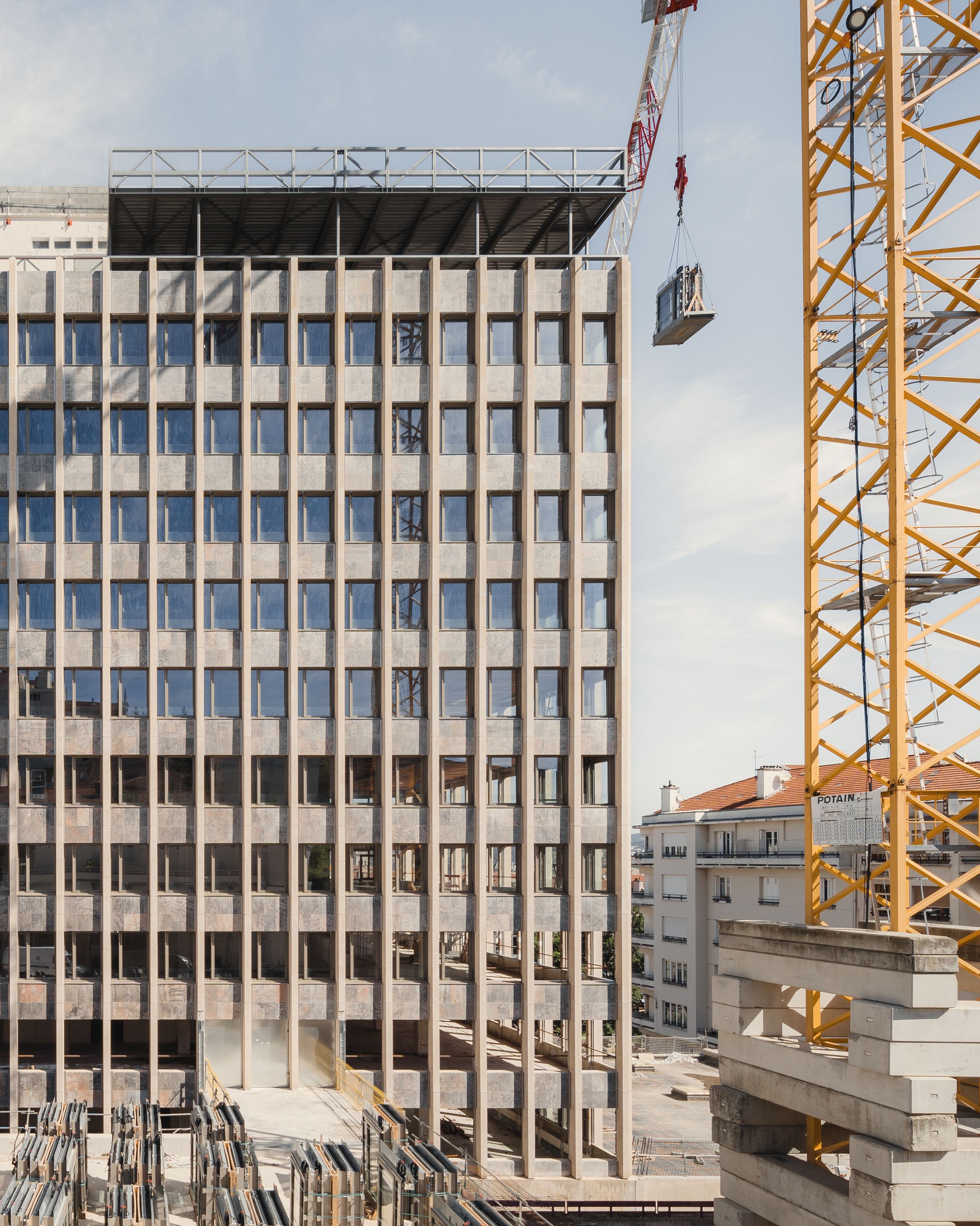 Chantier de réhabilitation de l’IGH Émile Loubet à Saint-Étienne : façade administrative des années 1970 en béton rythmé, curée, avec grue et éléments préfabriqués au sol.