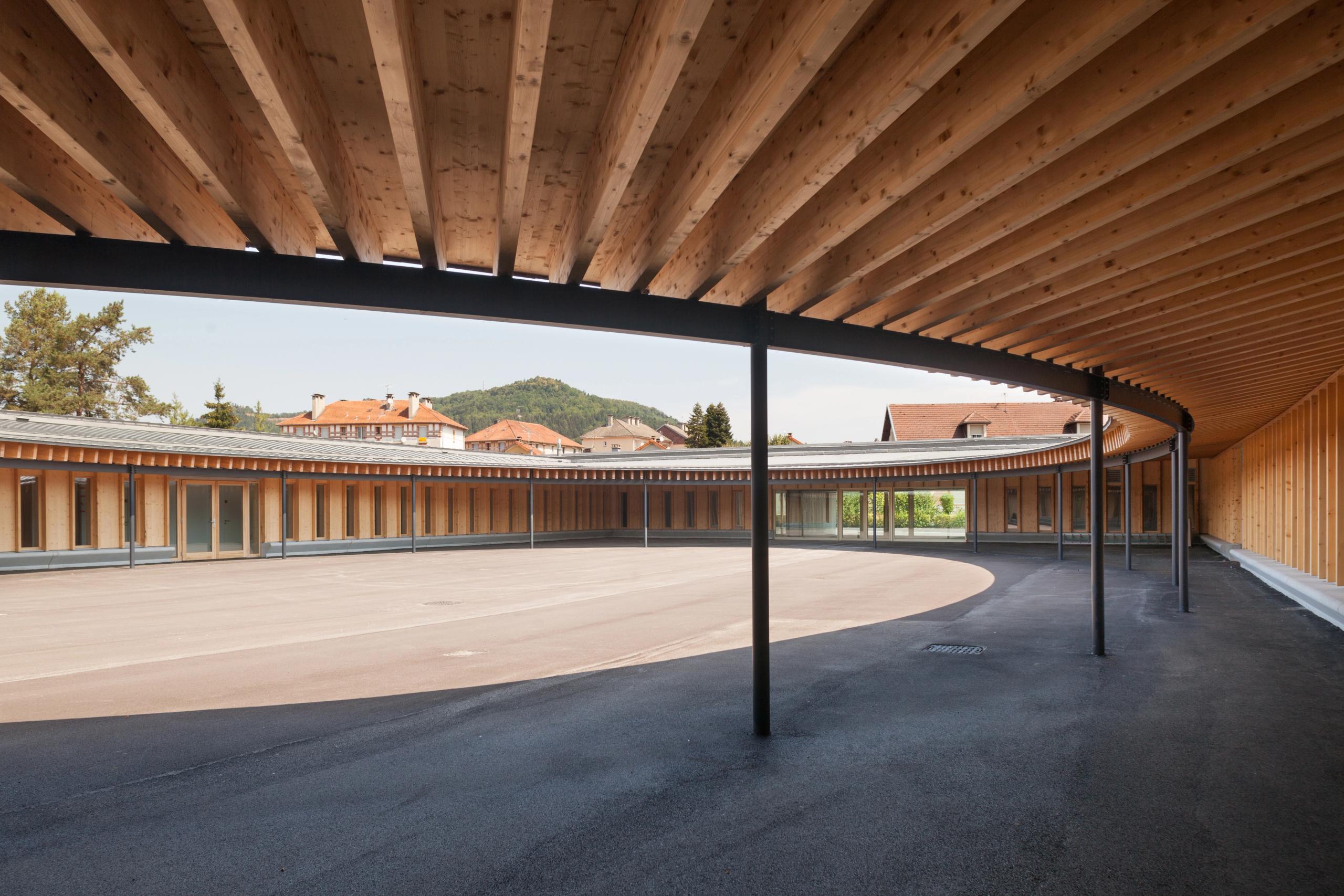 Cour elliptique couverte de l’école élémentaire Hubert Reeves à Champagnole, ossature bois apparente, poteaux métalliques et larges baies vitrées sur le paysage.