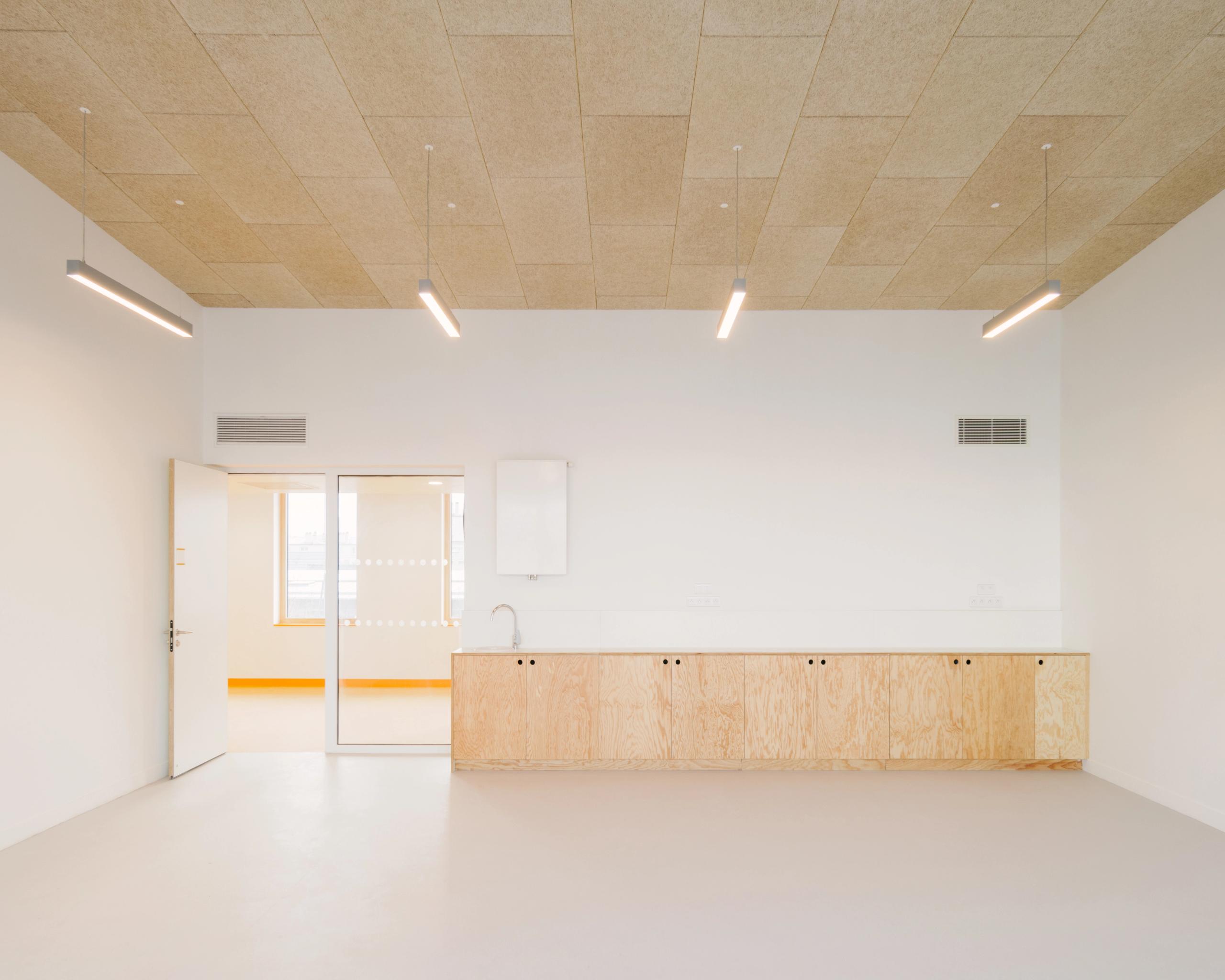 Intérieur d’une salle des groupes scolaires Jacqueline Auriol et Jean Jaurès au Bourget, avec plafond acoustique, éclairage linéaire et mobilier bas en bois clair.