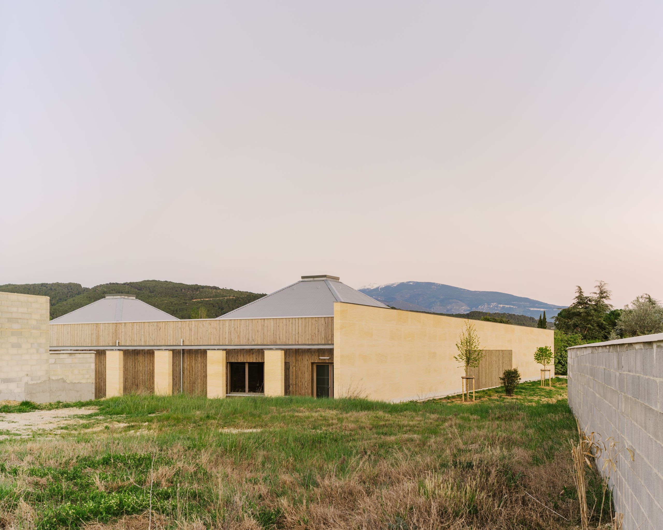 Pôle culture et jeunesse à Vaison-la-Romaine : volume bas en pierre massive et bois, toitures métalliques émergentes, implanté en lisière de lotissement périurbain.