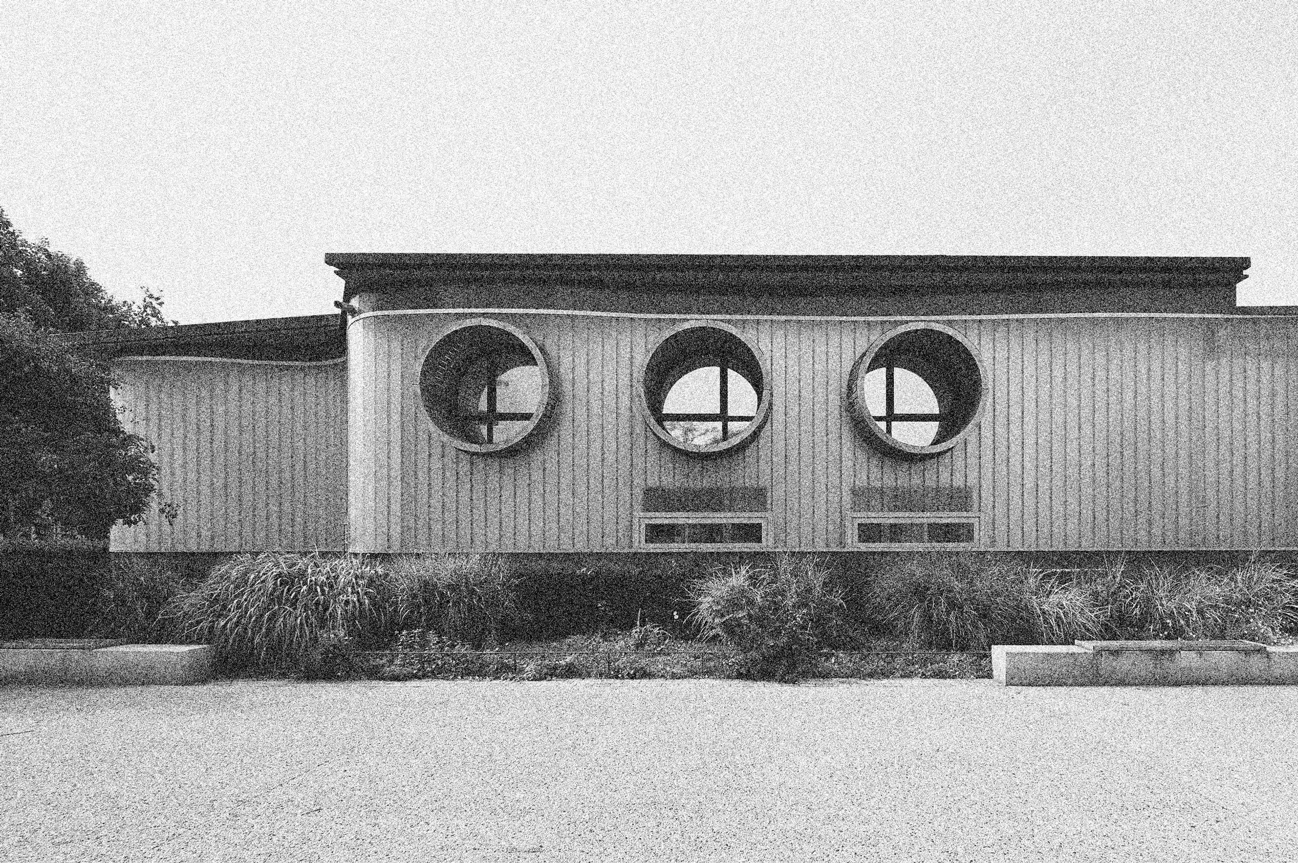 Document de processus du conservatoire d’Annemasse : façade existante à trame verticale, rythmée par trois baies circulaires, devant un parterre planté.