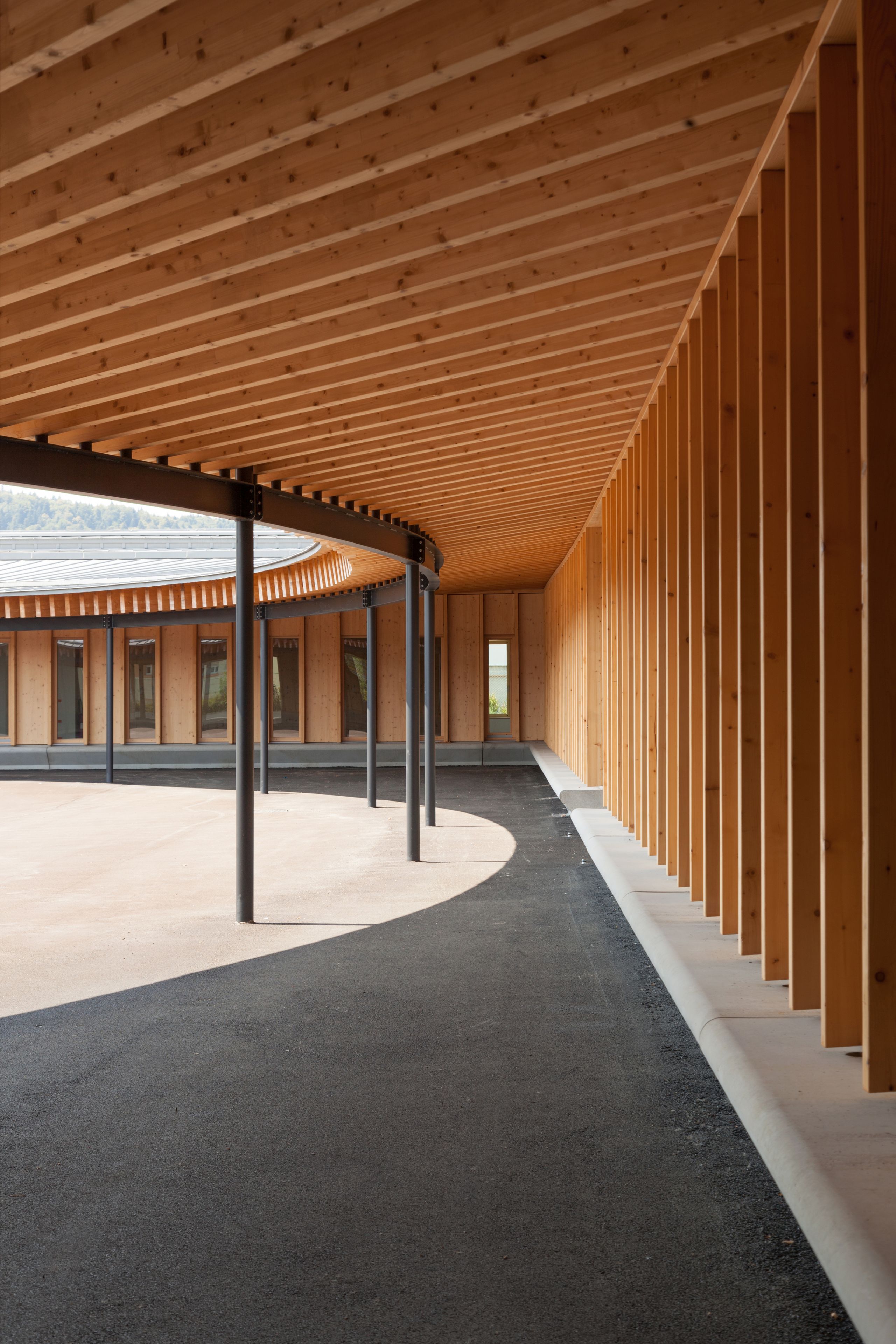 Galerie couverte elliptique de l’école élémentaire Hubert Reeves à Champagnole, structure bois apparente, poteaux métalliques et cour centrale ouverte sur le paysage.