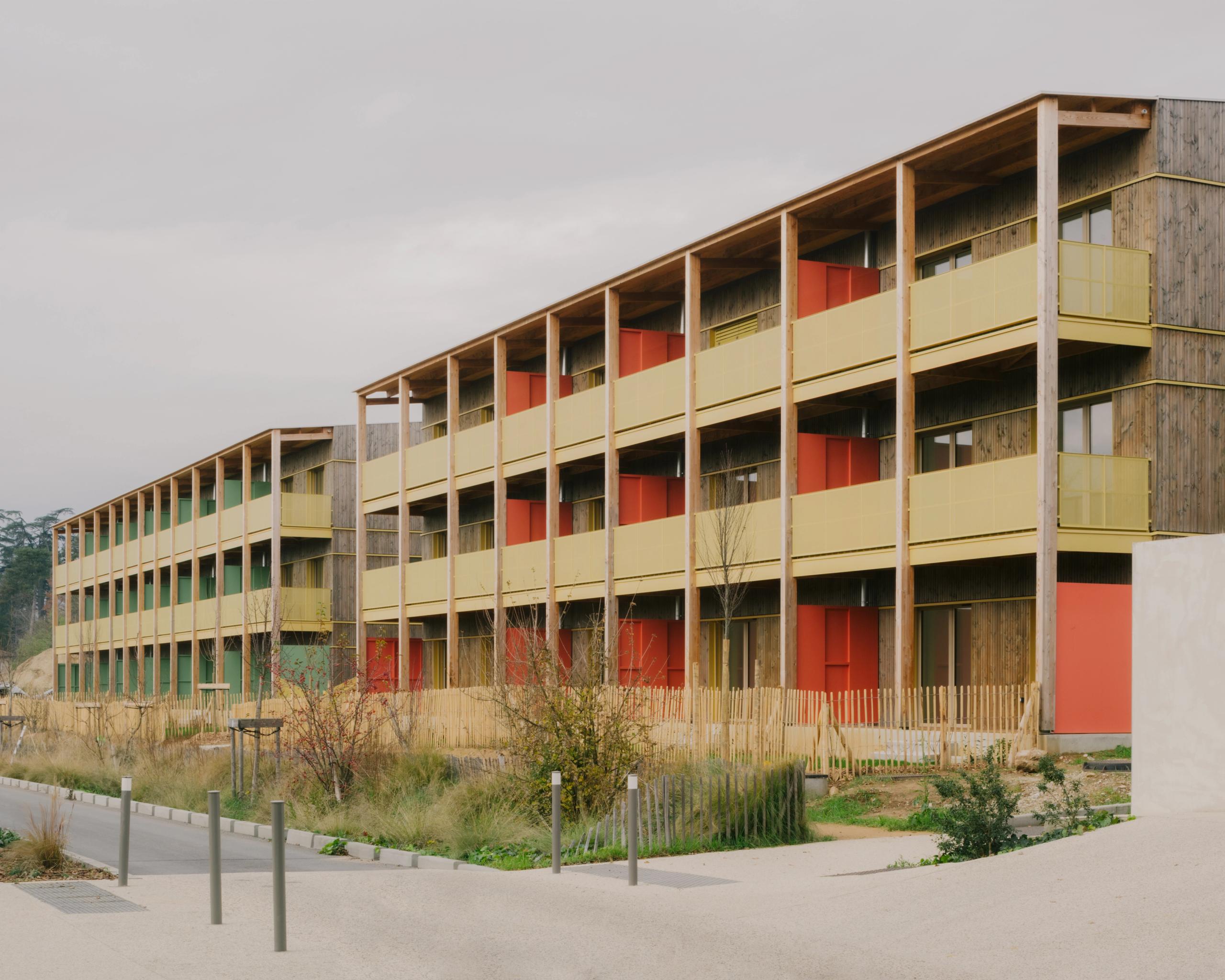 Vue des 84 logements à Trévoux par Tectoniques : volumes en gradins dans la pente, structure bois hybride, coursives extérieures et balcons colorés sur parc.