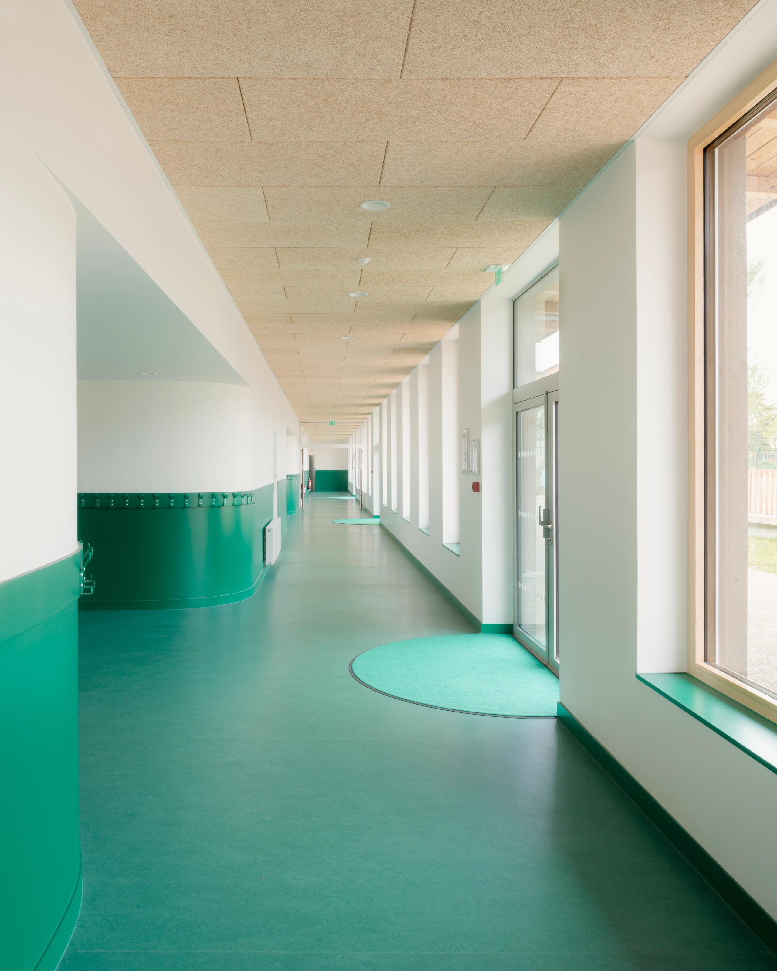 Couloir des groupes scolaires Jacqueline Auriol et Jean Jaurès au Bourget, sol et soubassements verts, plafond acoustique bois et façade vitrée sur cour.