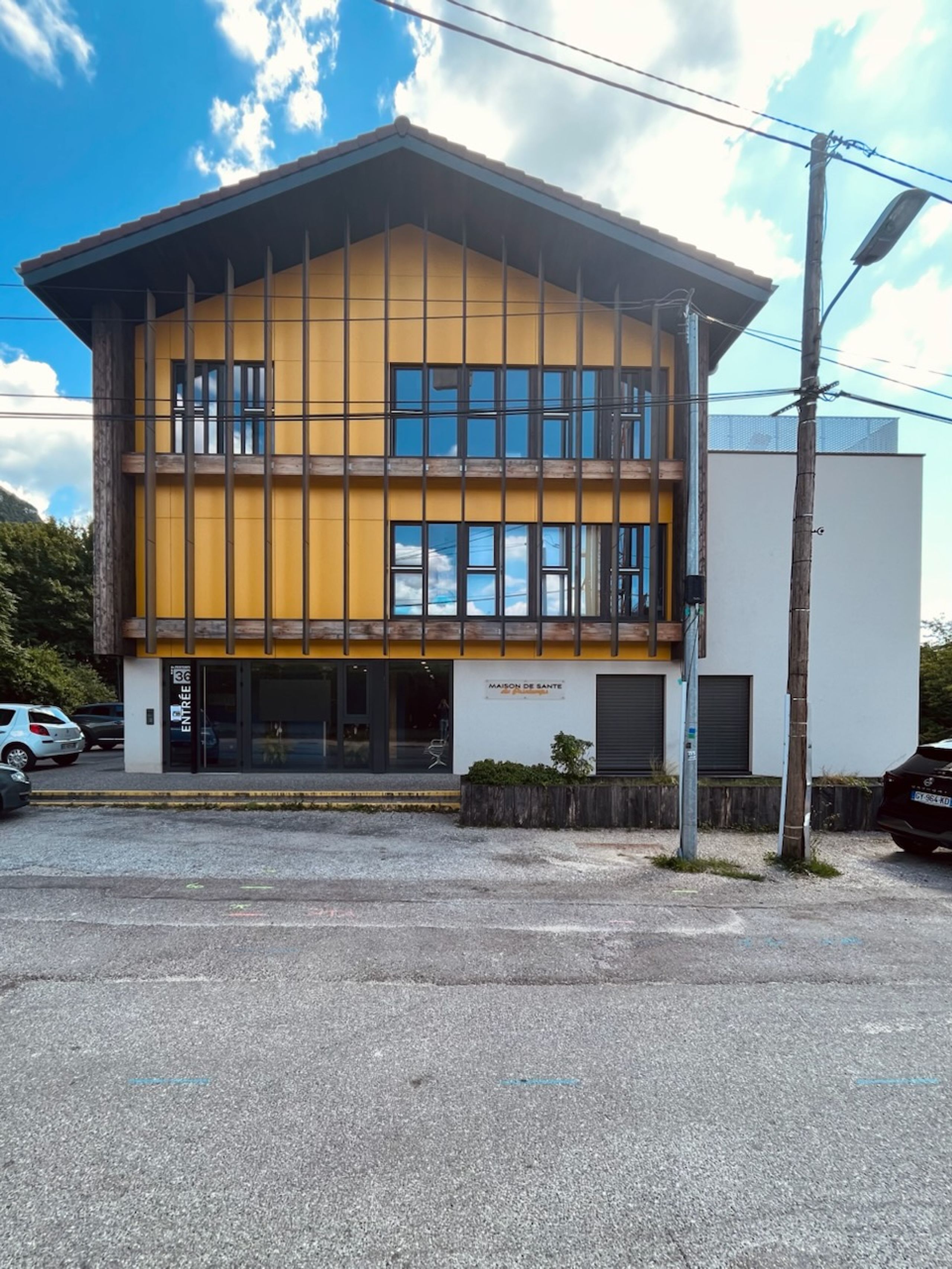 Façade de la maison de santé à Annecy, volume principal bois et enduit ocre avec trame verticale, larges baies vitrées et extension blanche en second plan.