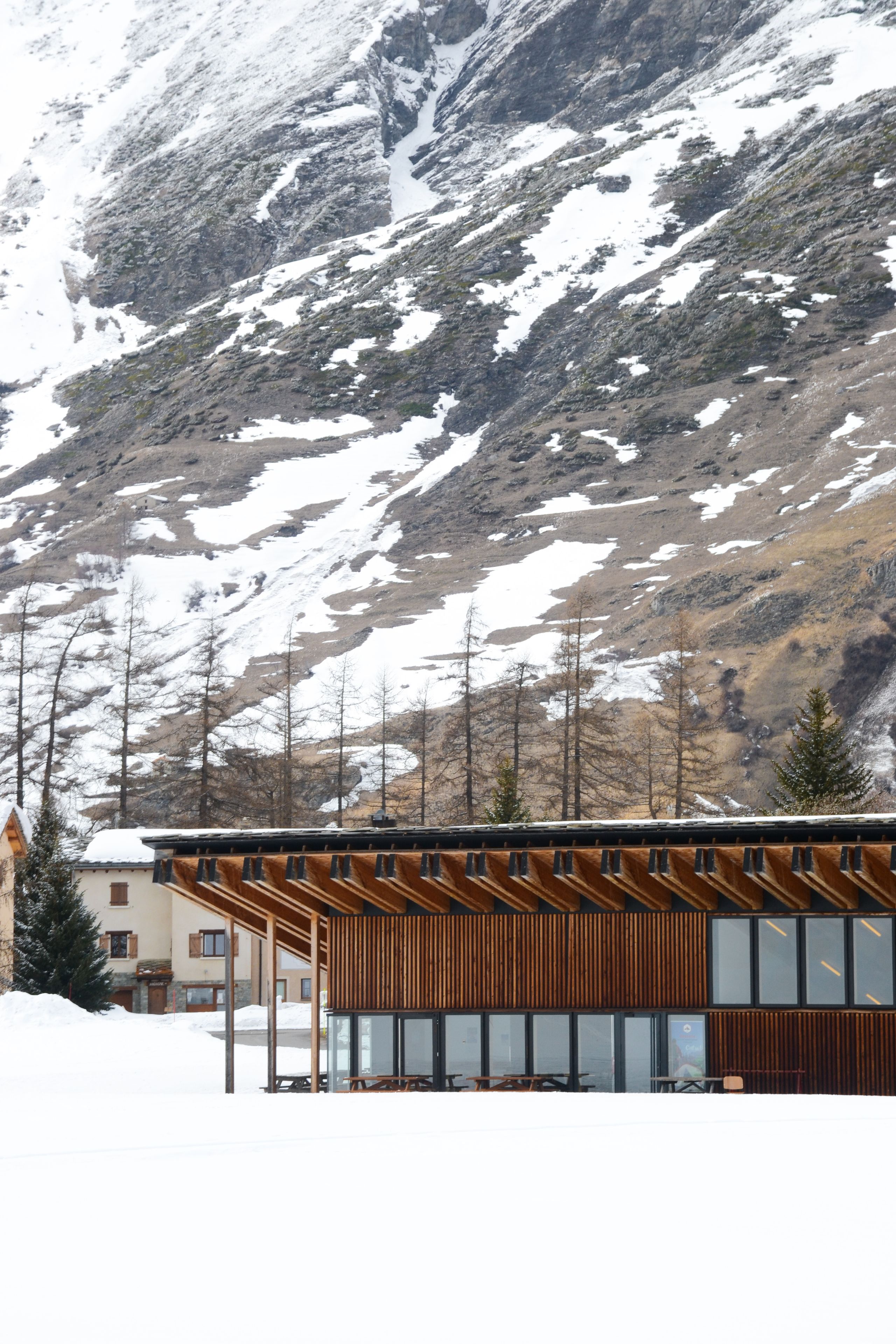 Espace nordique de Bessans : volume en bois à portiques apparents et façade vitrée, posé sur le manteau neigeux au pied d’un versant alpin minéral.