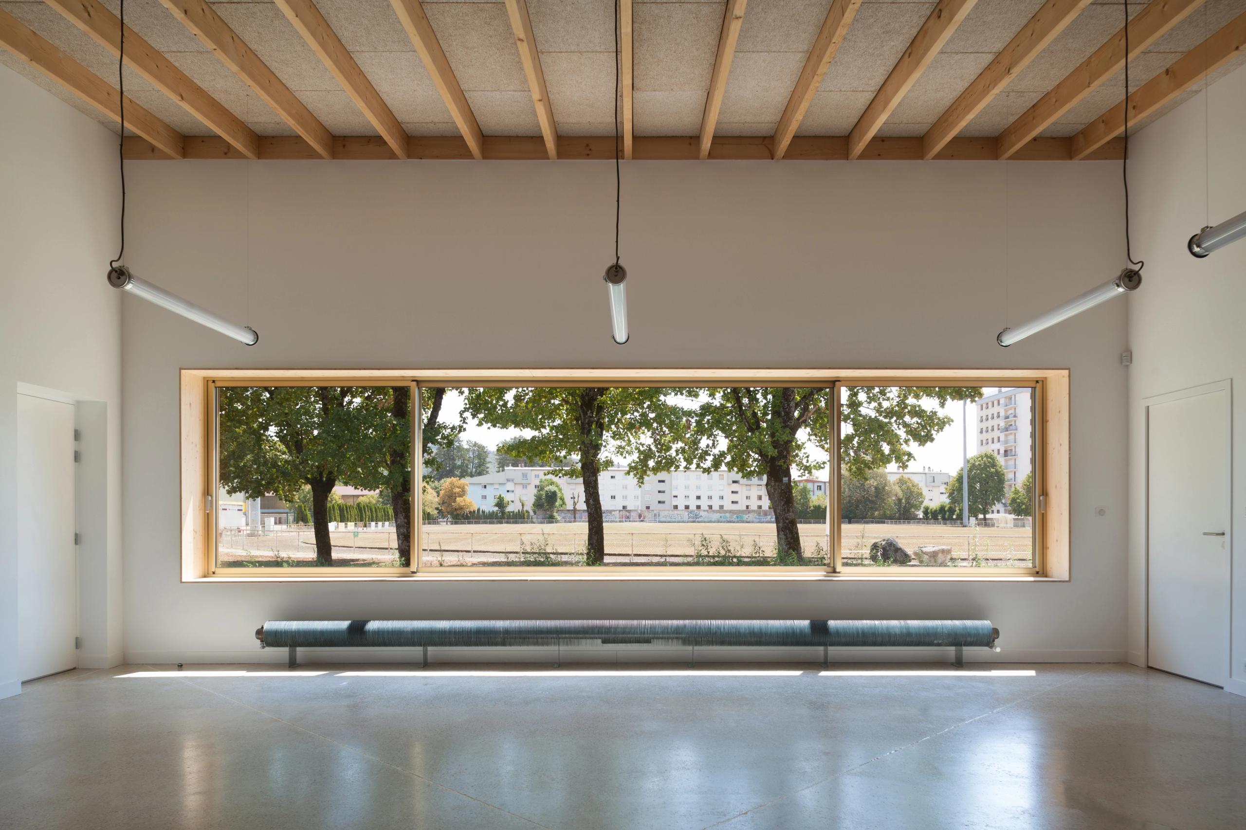 Intérieur d’une salle de l’école élémentaire Hubert Reeves à Champagnole, ossature bois apparente, grande baie horizontale cadrant la cour plantée.