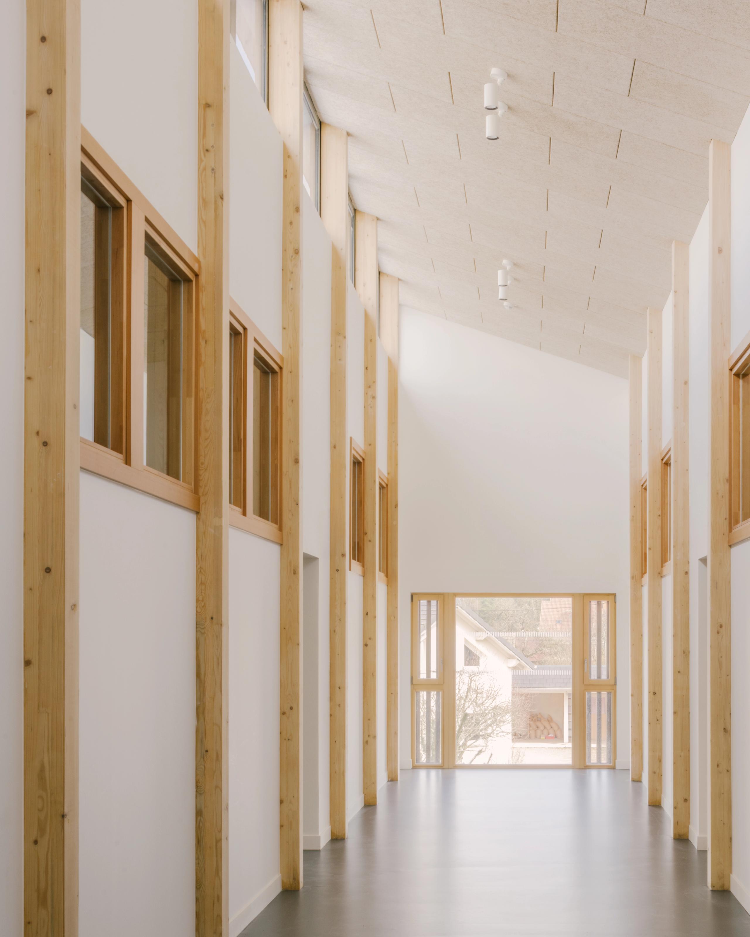 Rue intérieure du Collège du Ban à Vagney, galerie longitudinale en structure bois vosgien, baies vitrées en bout de perspective sur le paysage bâti.