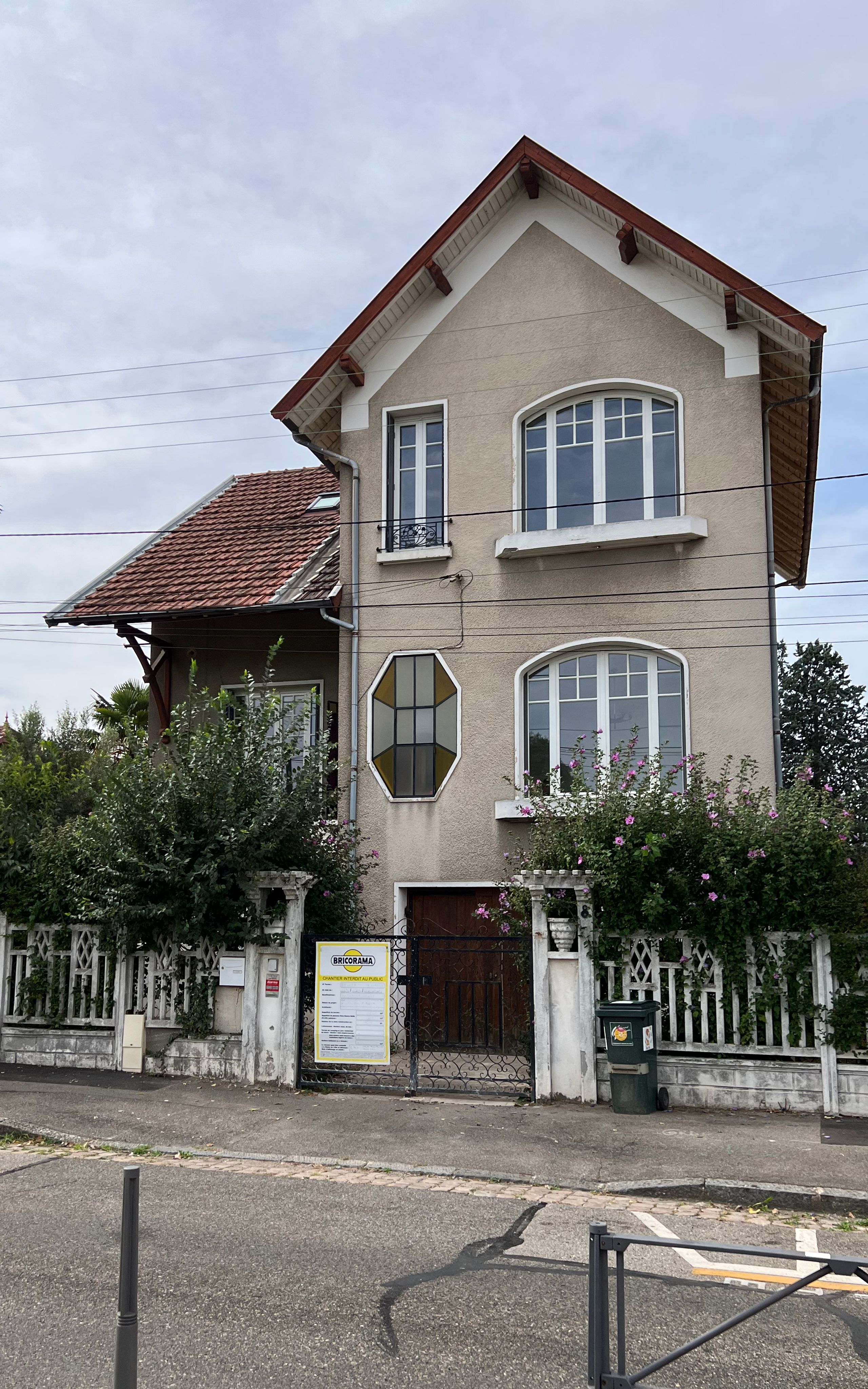 Façade sur rue d’une maison faubourienne à Lyon, volume enduit R+2 sous toiture tuiles, baies cintrées et octogonale, clôture maçonnée et végétation dense.