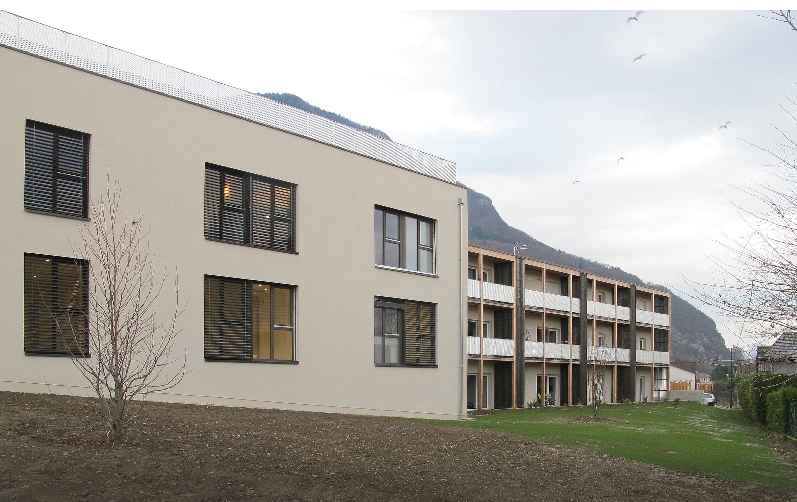 Vue de la maison de santé à Annecy, volume enduit sobre et ailes à balcons filants en structure bois, grandes baies vitrées ouvrant sur un jardin au pied de la montagne.