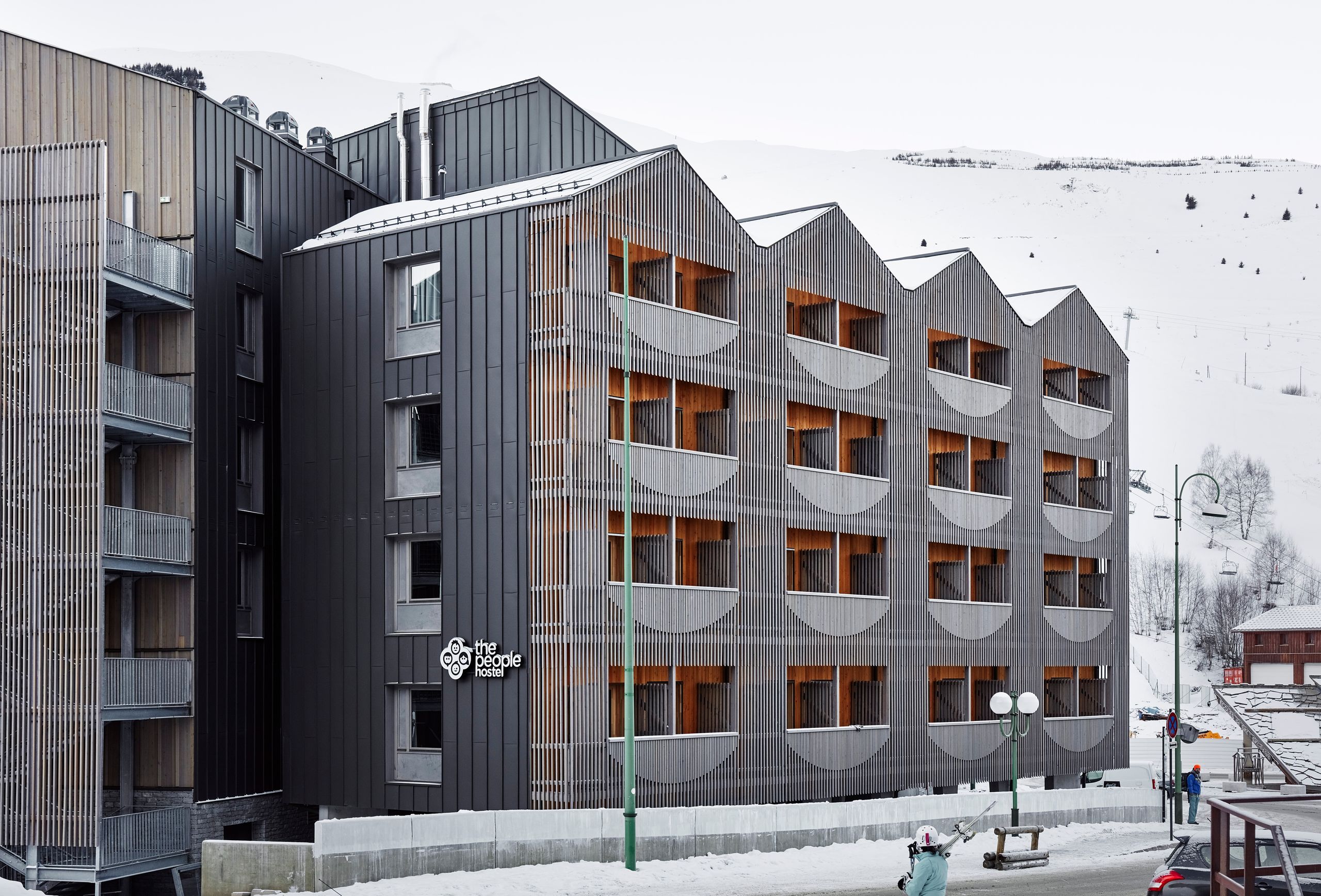 Façade de l’auberge de jeunesse The People Hostel aux Deux Alpes, volume fragmenté en métal sombre et double peau bois à claire-voie avec balcons semi-circulaires.