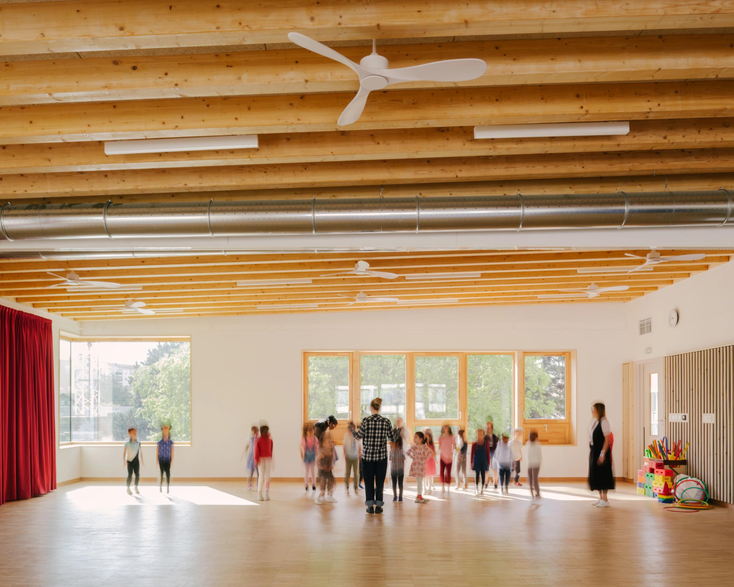 Intérieur lumineux du Groupe scolaire Kennedy à Lyon : grande salle en bois avec plafond à poutres apparentes, gaines métalliques et larges baies sur la cour arborée.