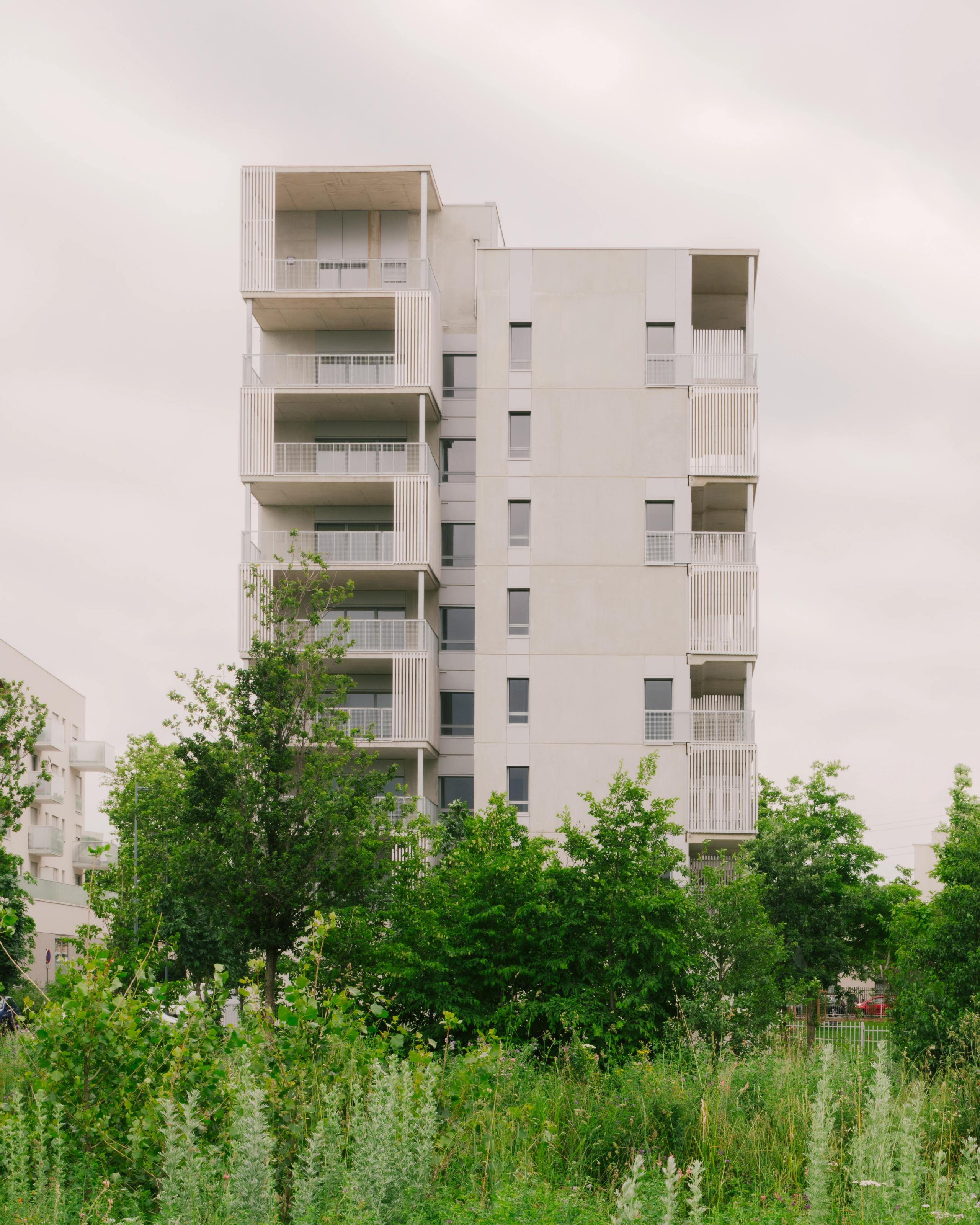 Vue du bâtiment de 116 logements à Vénissieux, volume fragmenté en plots avec balcons filants en exosquelette métallique, sur socle paysager planté.