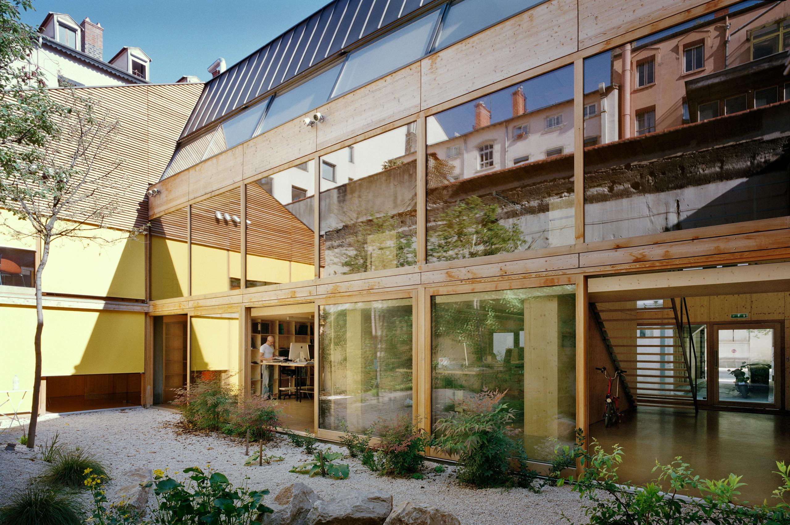 Réhabilitation d’un local d’activité à Lyon par Tectoniques : façade patio vitrée et structure bois apparente, jardin minéral central éclairant les espaces de travail.
