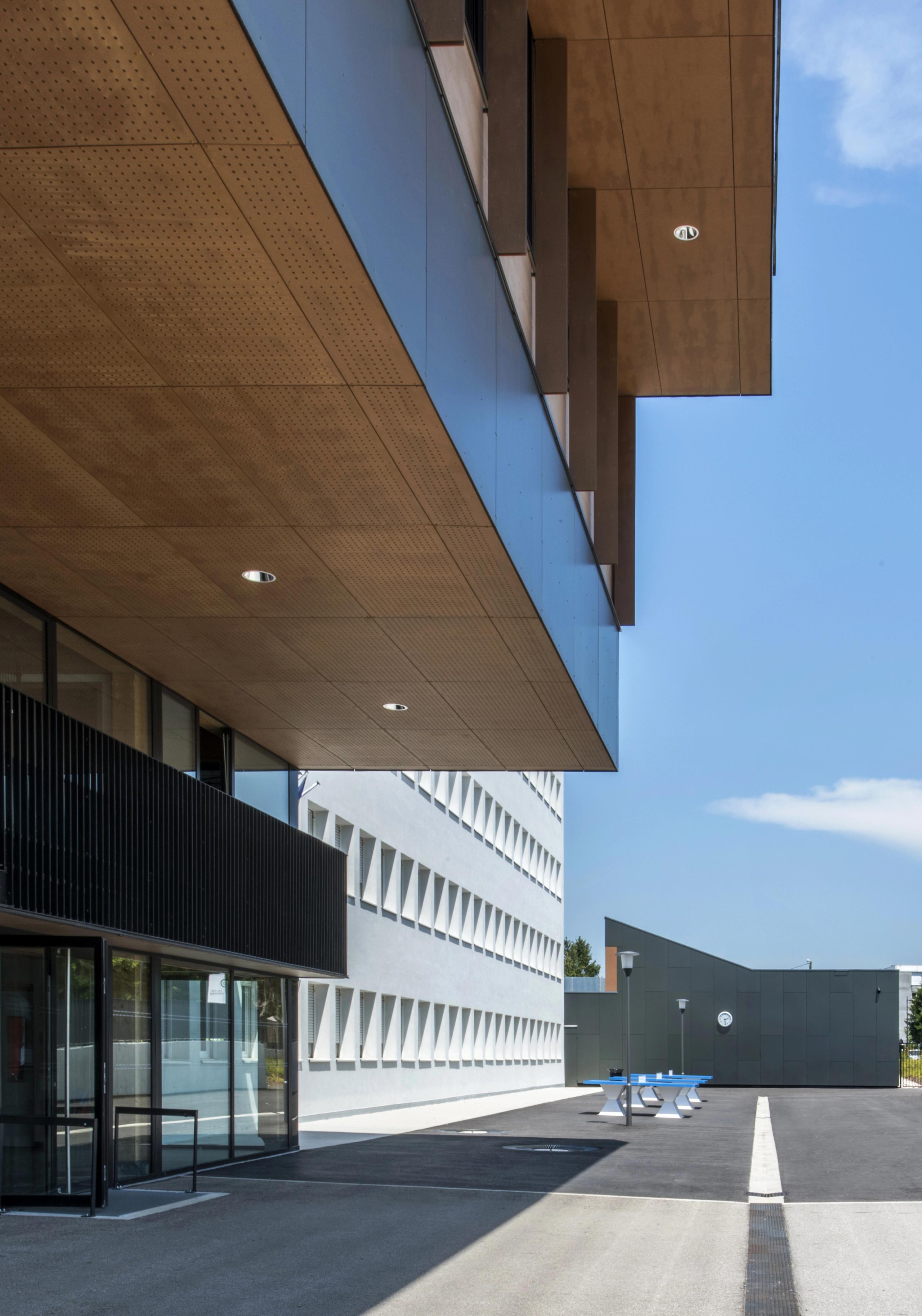 Façade restructurée du collège Pierre-Joseph Proudhon à Besançon, galerie couverte en bois, volume blanc percé de baies triangulaires et cour minérale ensoleillée.