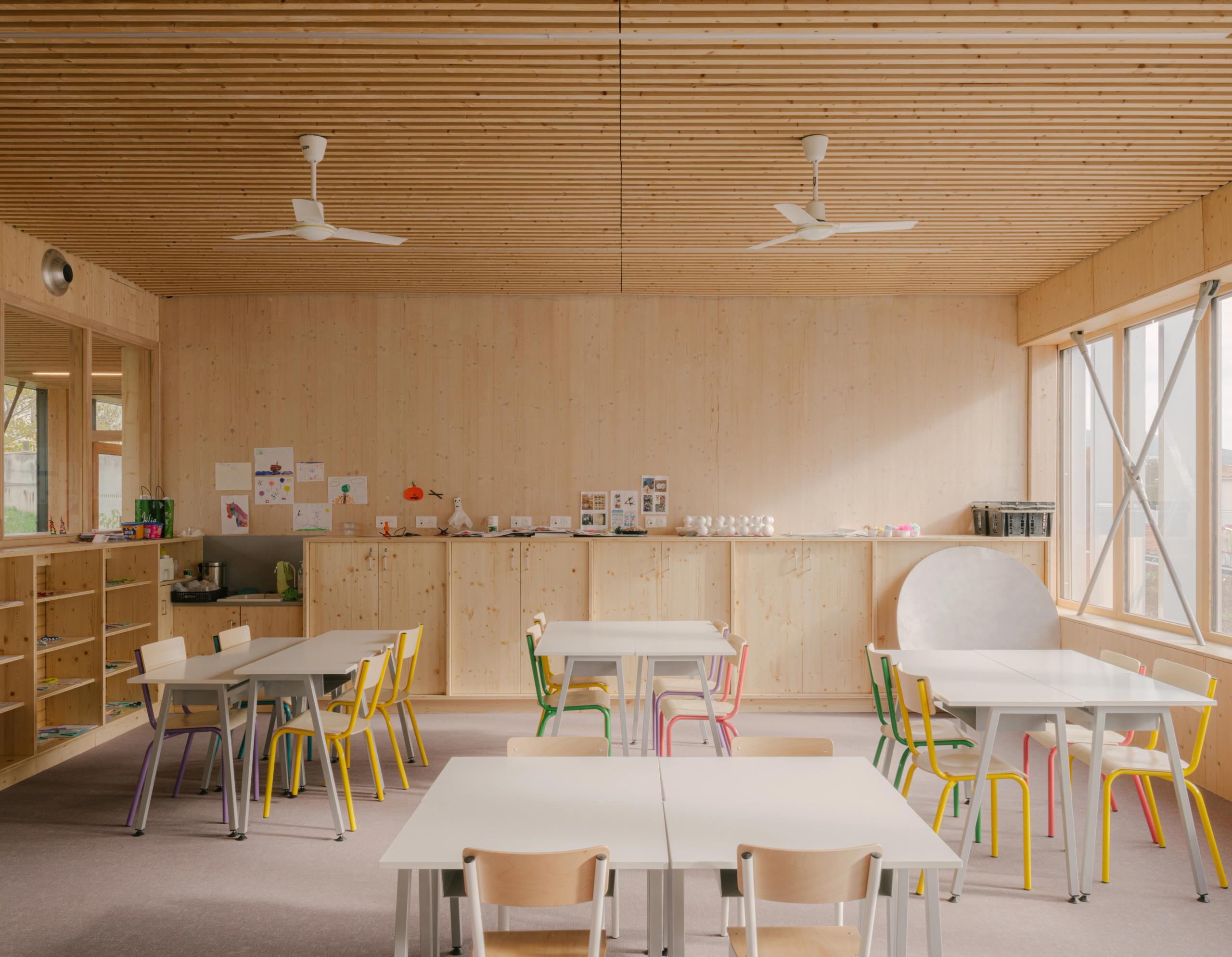 Salle de classe du groupe scolaire Claudie Haigneré aux Pennes-Mirabeau, structure bois apparente, plafond à lames, grandes baies vitrées et mobilier coloré.