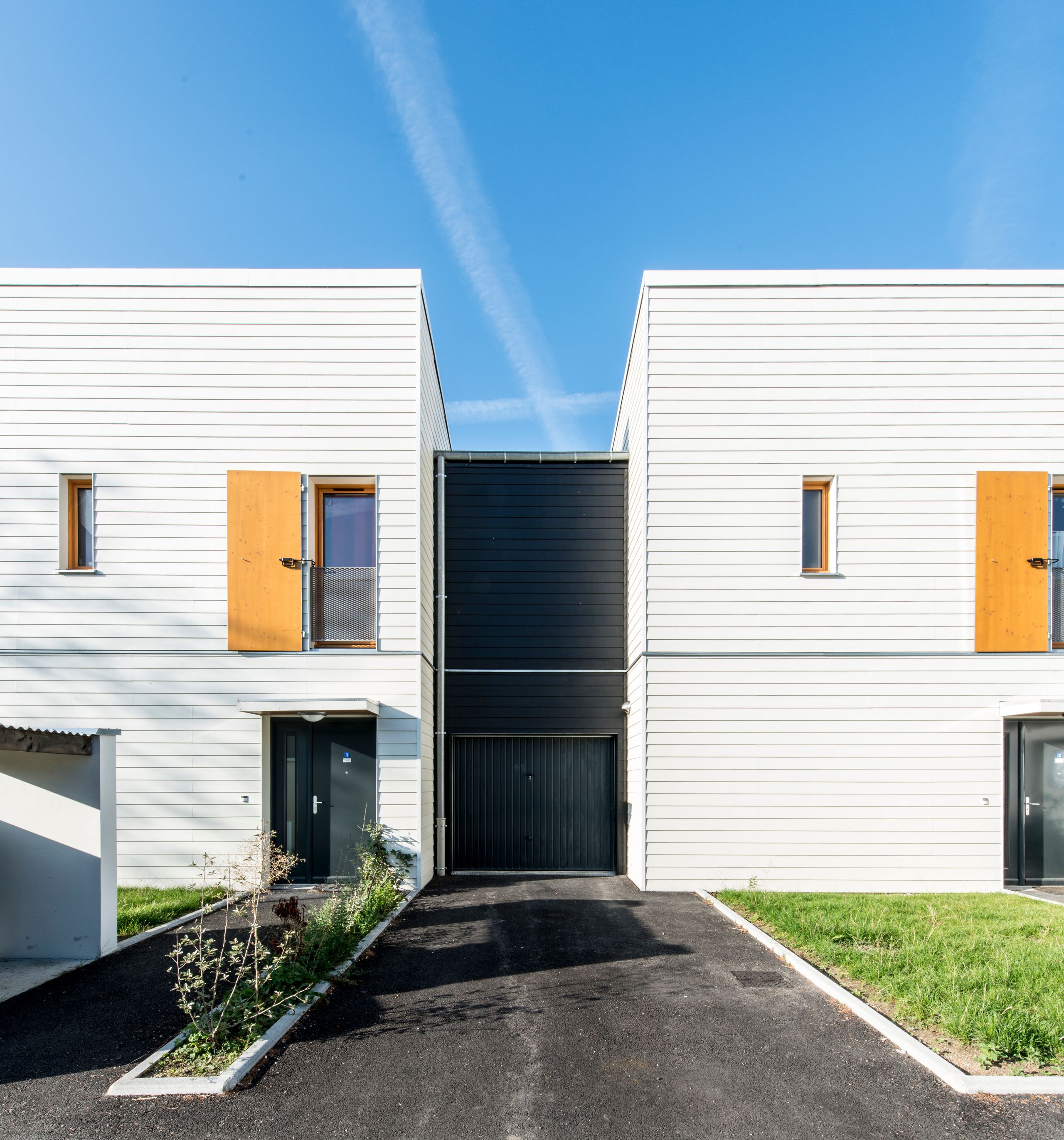 Façade de logements 5D à Rive-de-Gier par Tectoniques, volumes fragmentés en bardage clair et noir, volets bois, accès garage et chemin piéton paysager.