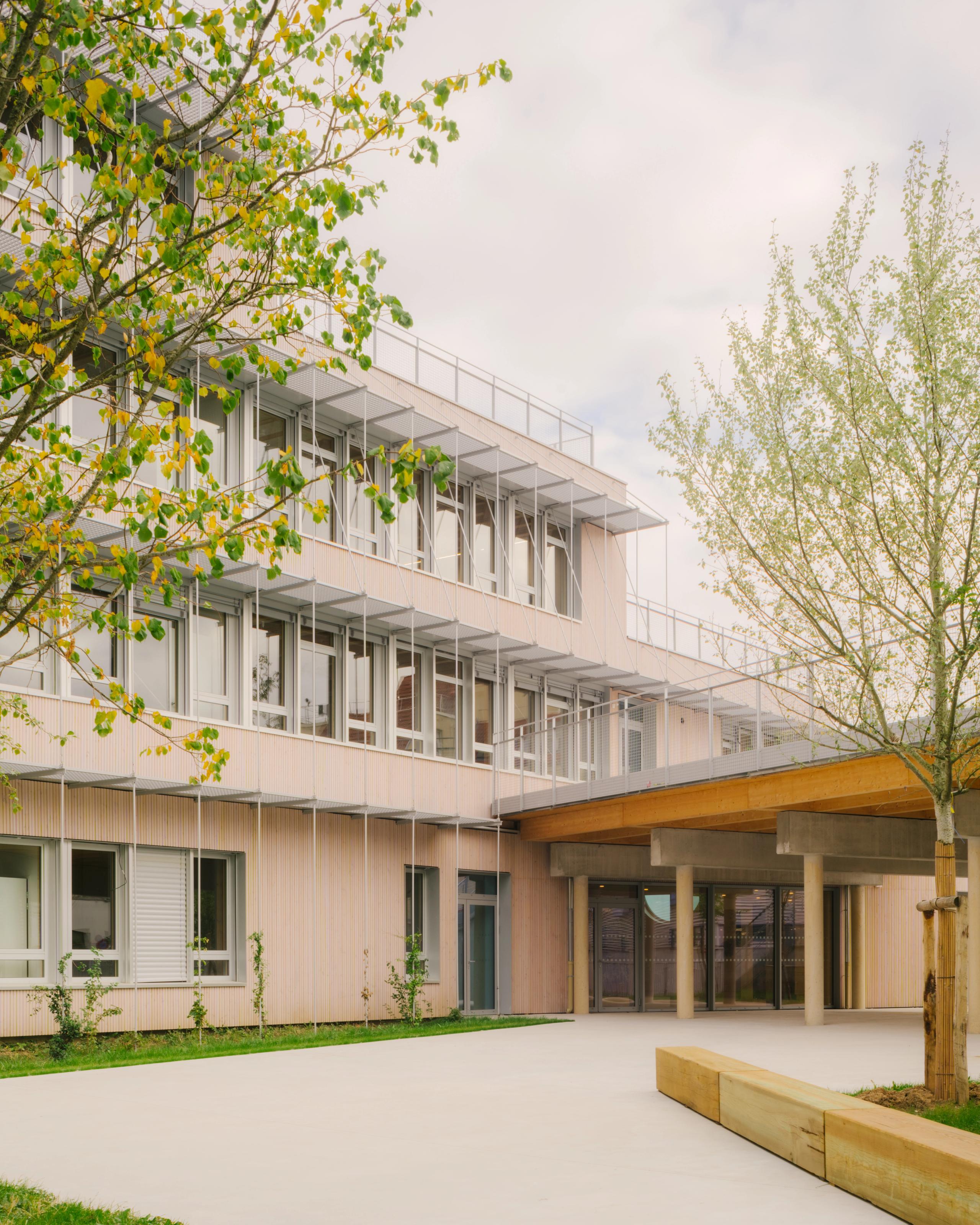 Façade bois et métal du Groupe scolaire Dominique Frelaut à Colombes, volumes en gradins, casquette d’entrée structure bois-béton, cour plantée en premier plan.