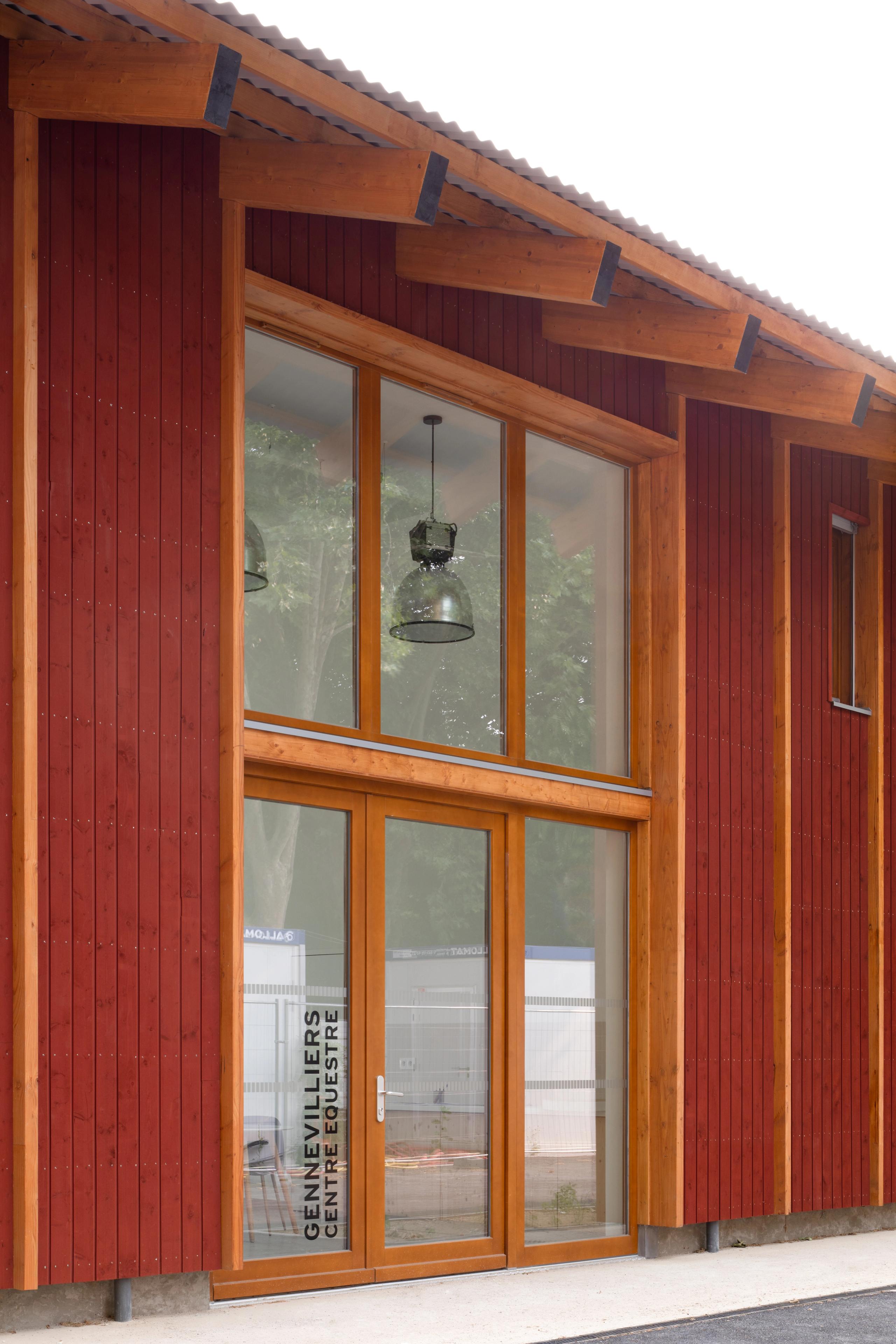 Timber façade of the Gennevilliers equestrian center with Falun red vertical boarding, large glazed entrance and exposed roof structure facing the street.