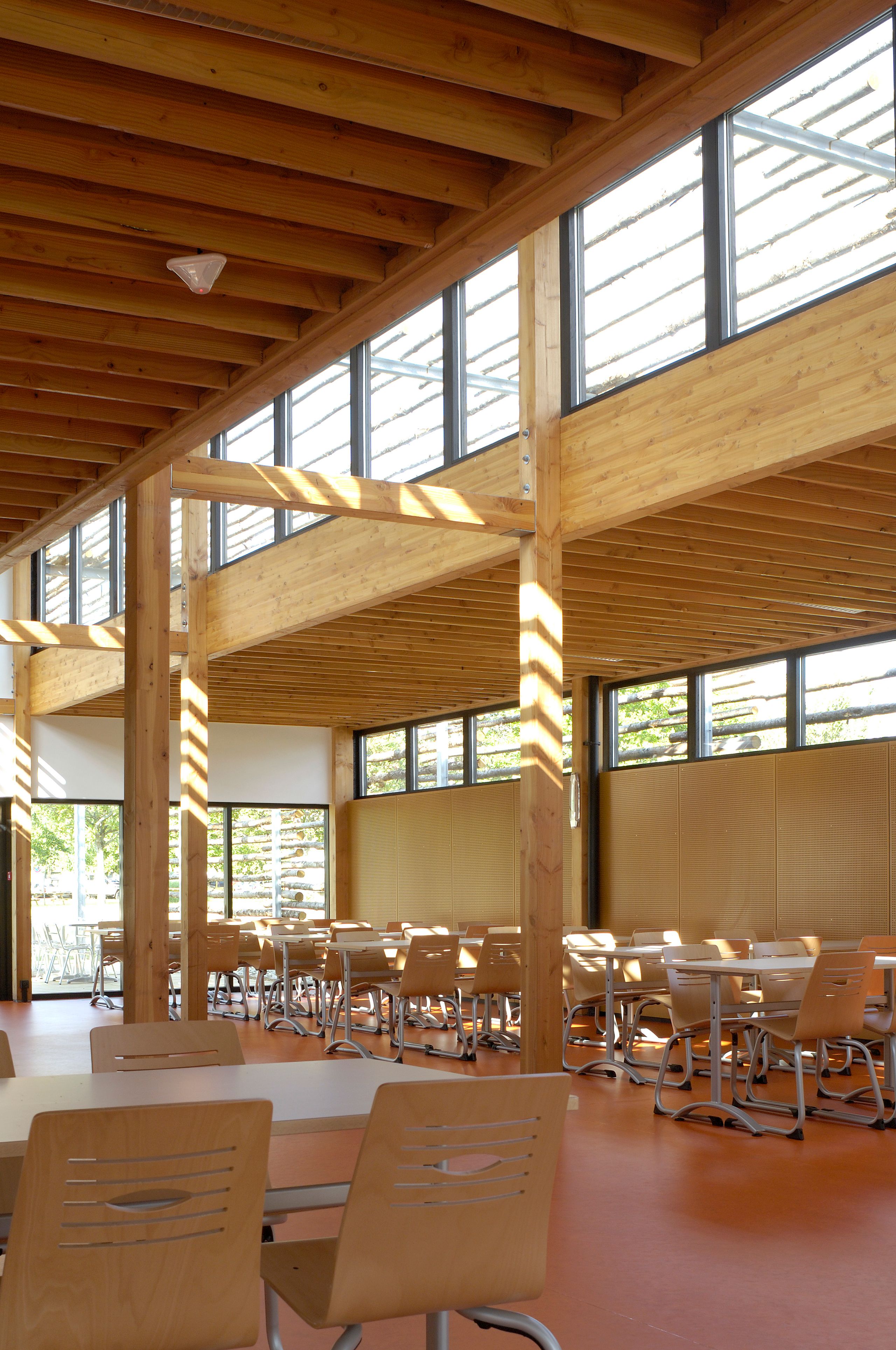 Intérieur du restaurant administratif du CETE à Bron : vaste salle à structure bois apparente, grandes baies vitrées, lumière zénithale et mobilier de réfectoire.