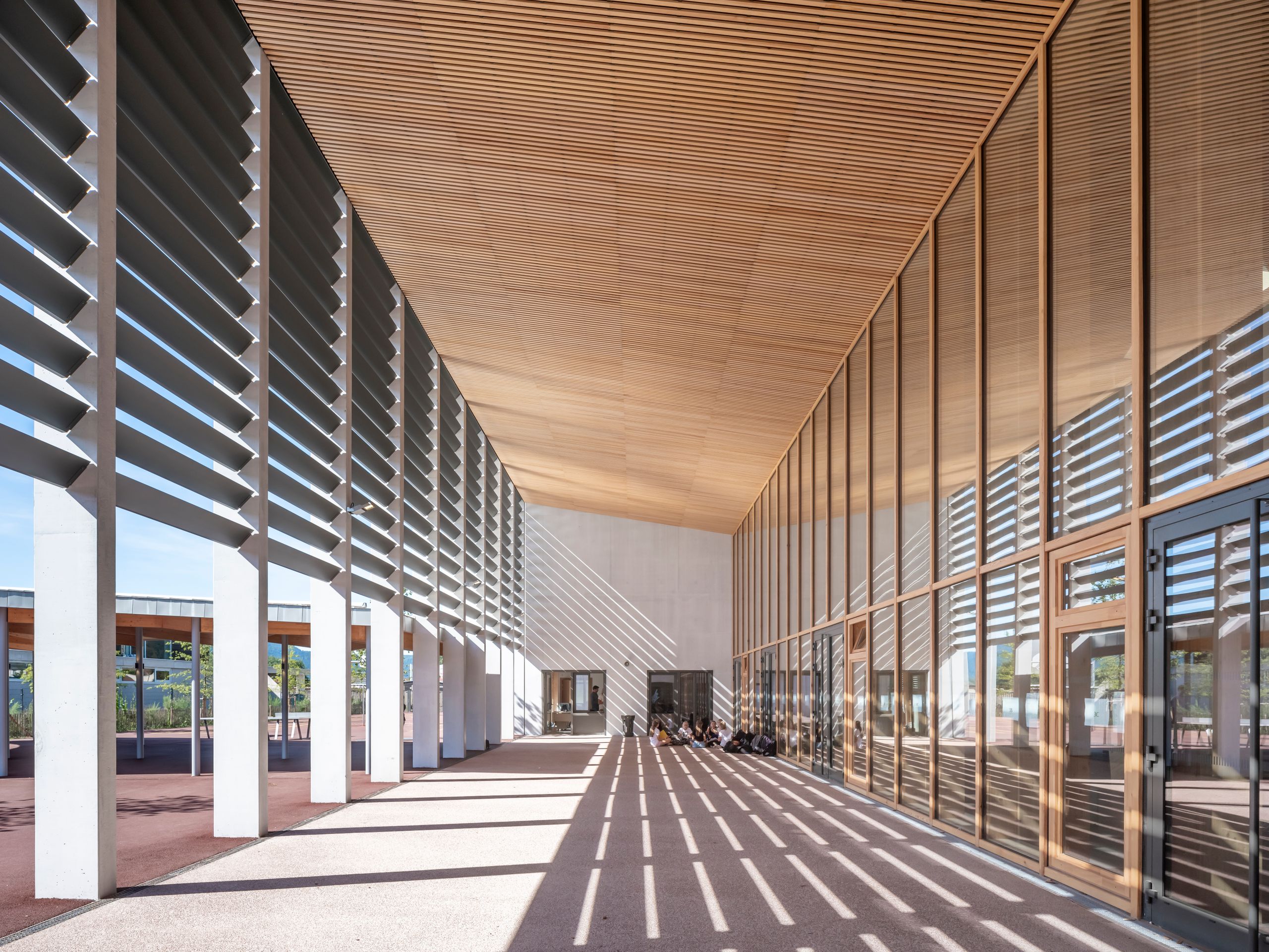 Vue du grand préau du Collège du Ban à Vagney : un portique monumental en béton et lames brise-soleil métalliques cadre un espace couvert ouvert sur la cour, tandis qu’une façade vitrée à ossature bois, en double hauteur, prolonge la rue intérieure et laisse filtrer la lumière, dessinant au sol un maillage régulier d’ombres portées.