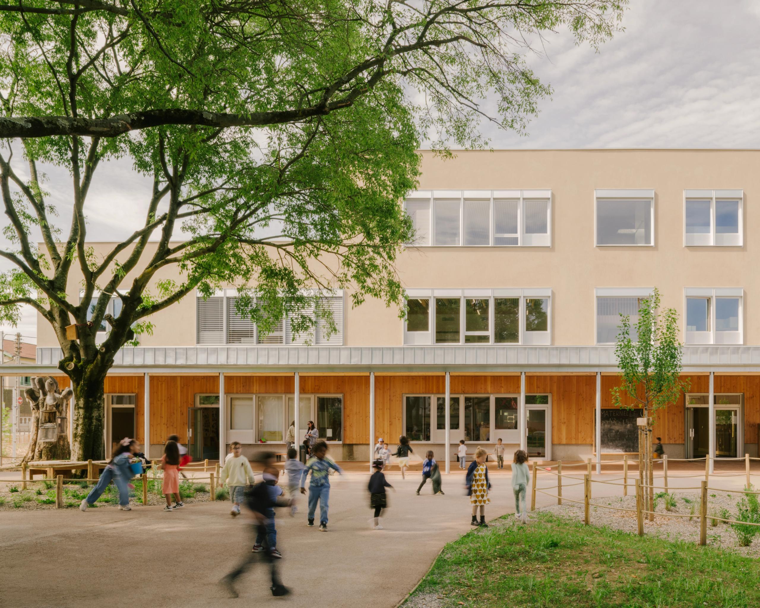Façade du groupe scolaire Kennedy à Lyon : volume simple enduit clair sur socle bois, préau sur cour de récréation végétalisée animée par les élèves.