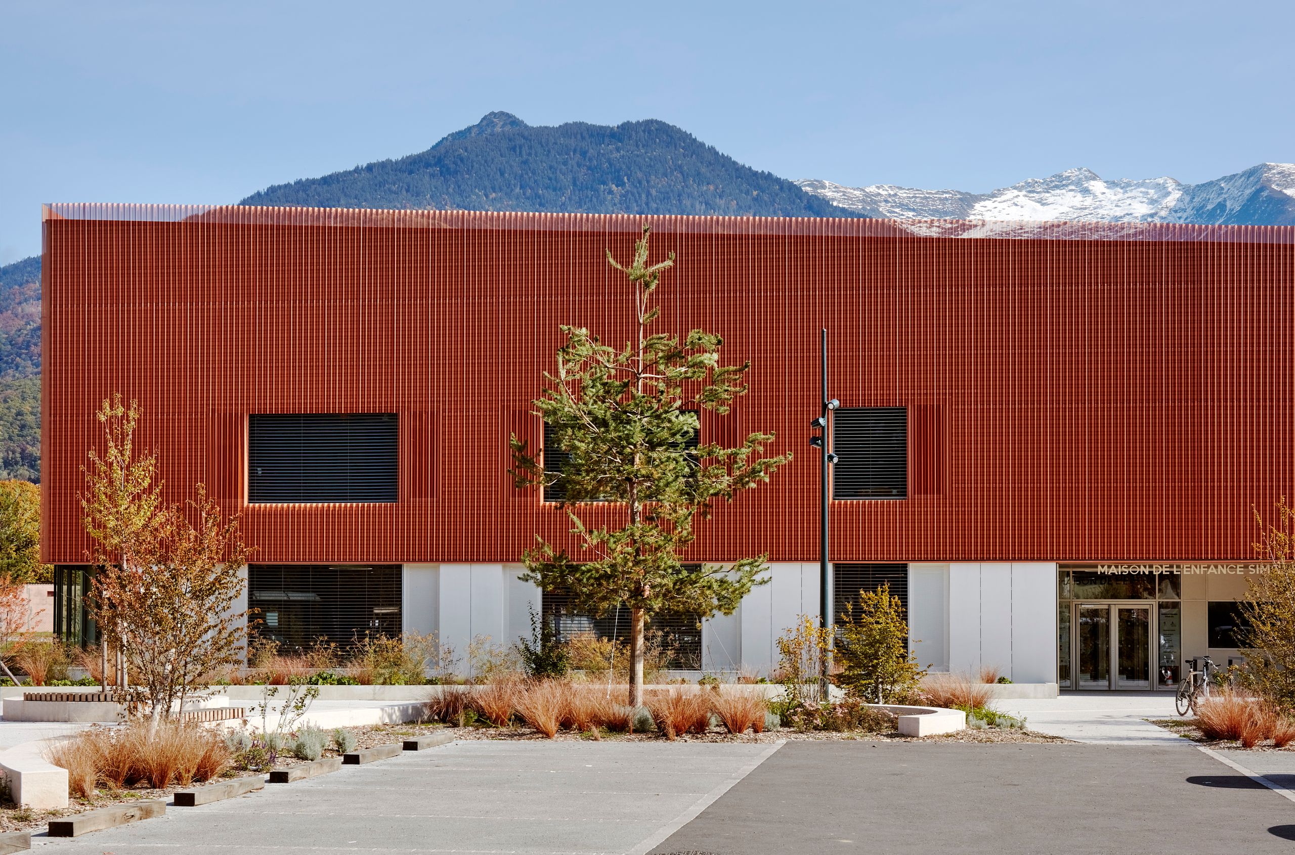 Façade compacte cuivrée de la Maison de l’enfance Simone Veil à Albertville, volume épais sur socle vitré, cadrant les massifs alpins en arrière-plan.