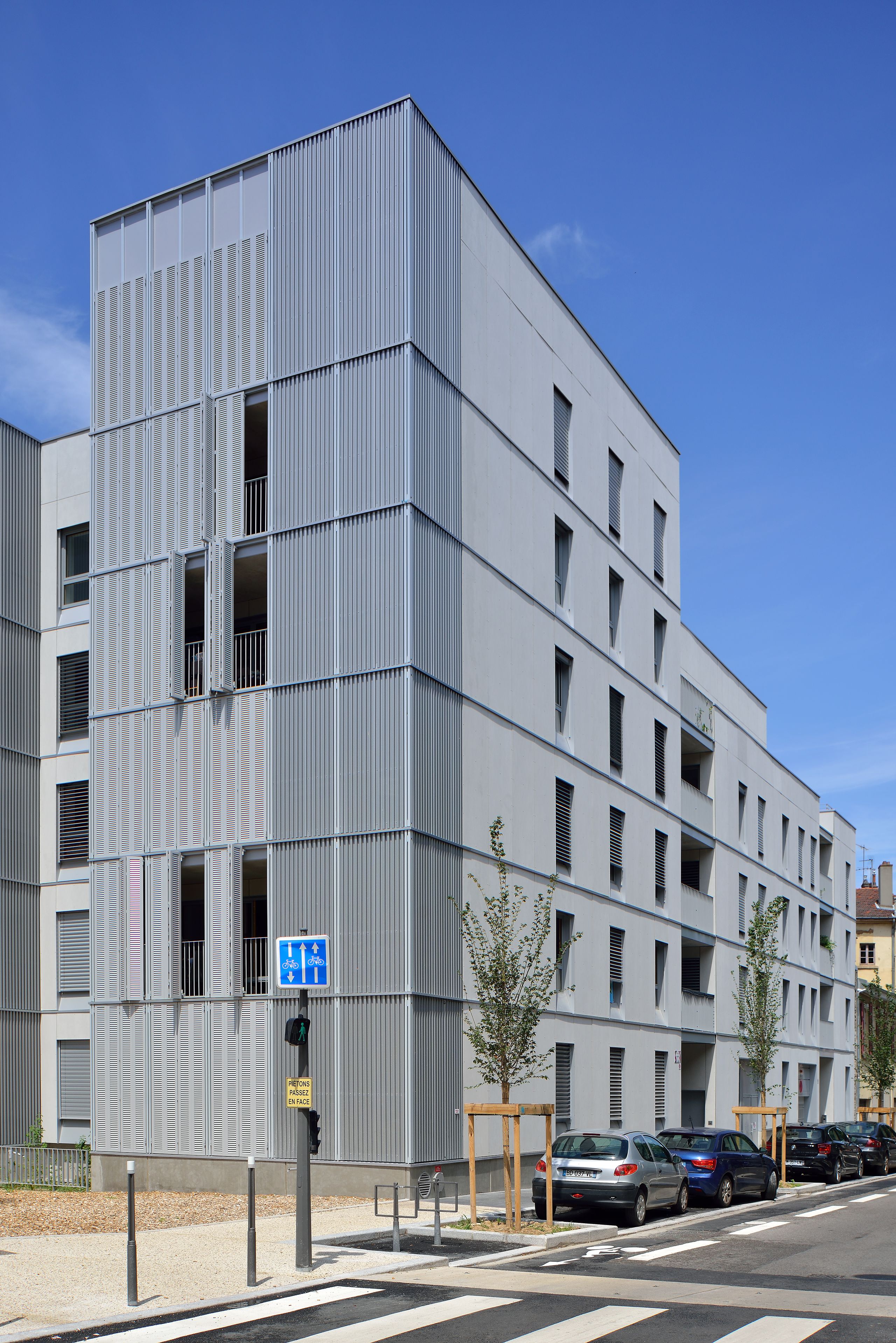 Façade de l’immeuble de 28 logements à Villeurbanne, volumes dégressifs, loggias en retrait et persiennes métalliques verticales sur rue apaisée.