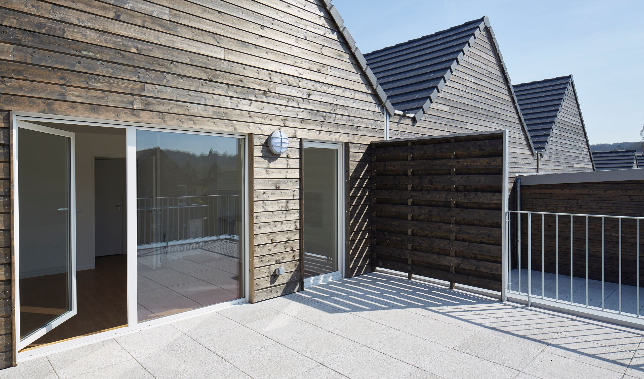 Terrasse des 36 logements à Vauhallan par Tectoniques, bardage bois à claire-voie, toitures à deux pans, grandes baies vitrées et garde-corps métalliques.