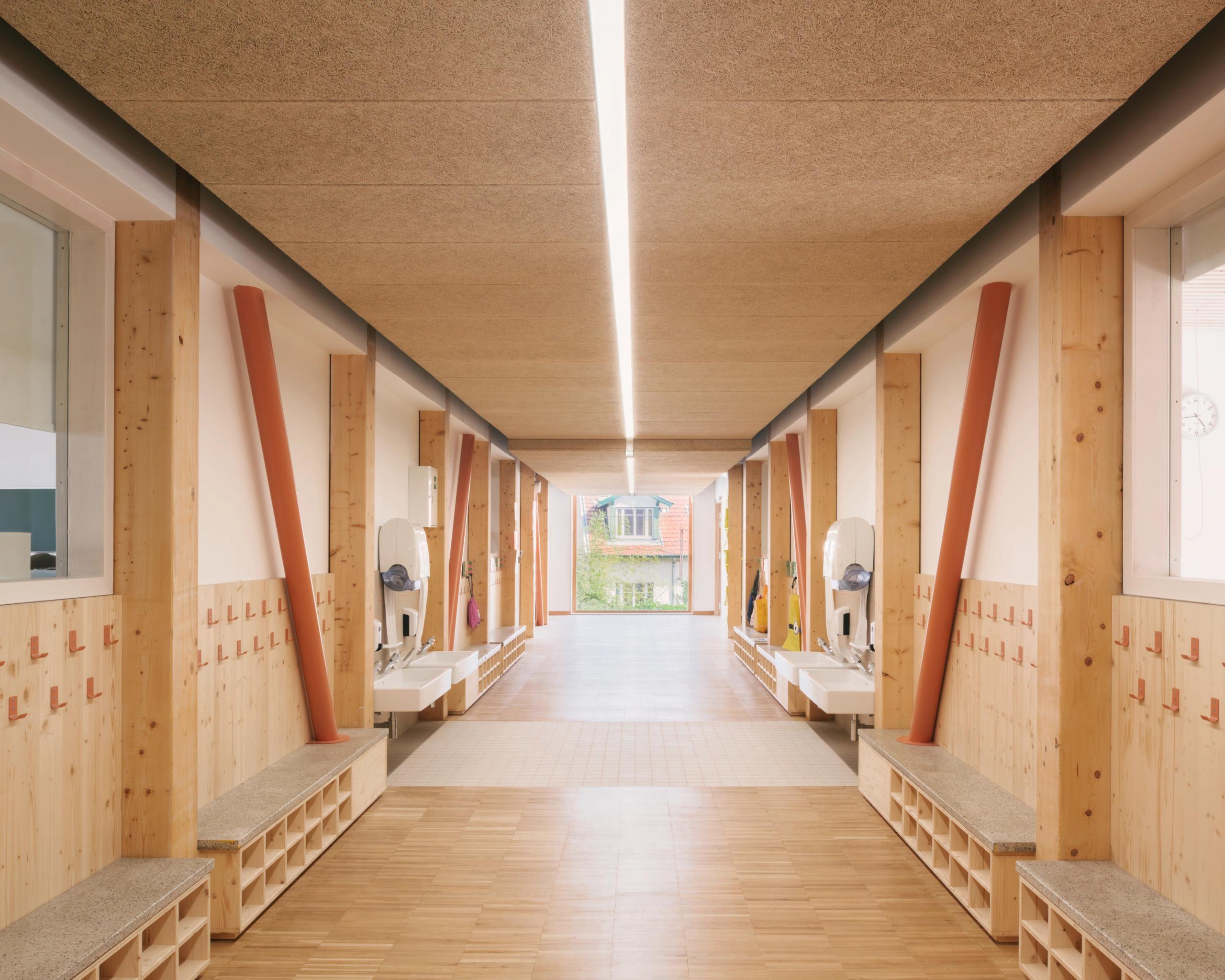 Couloir du groupe scolaire Kennedy à Lyon : structure bois apparente, poteaux inclinés, bancs et rangements intégrés, lumière naturelle en fond de perspective.