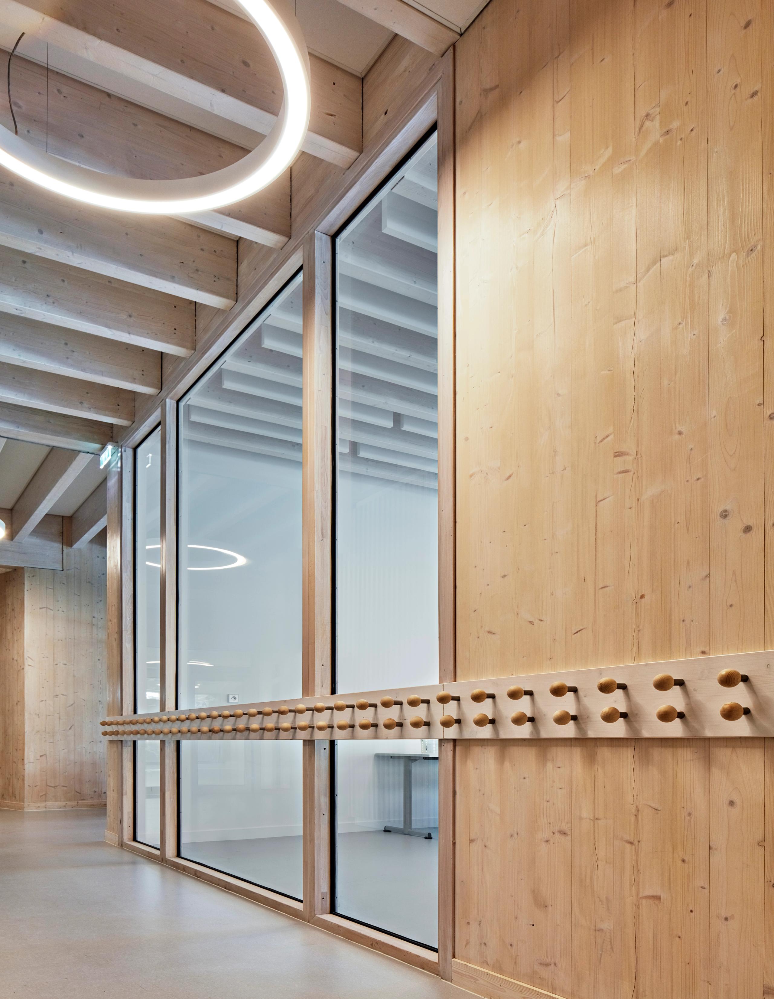 Intérieur en bois apparent de la Maison de l’enfance Simone Veil à Albertville, avec cloison vitrée, patères à hauteur d’enfant et éclairage circulaire suspendu.