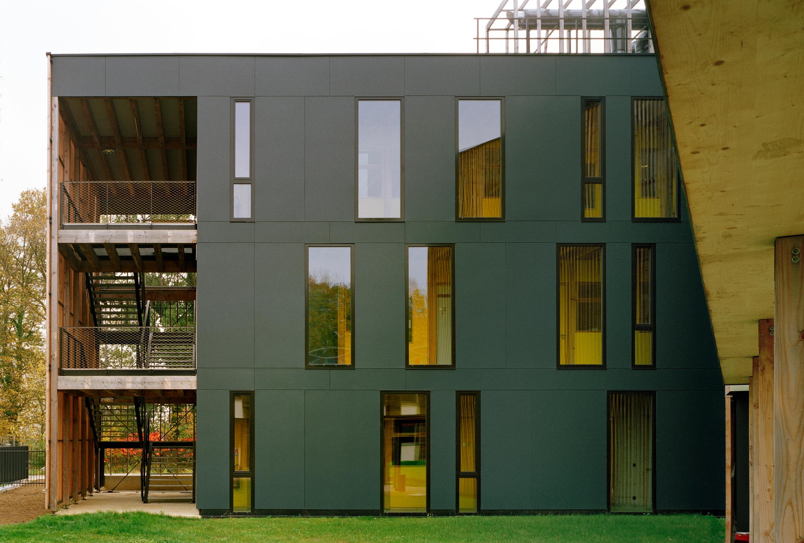 Façade du laboratoire INRAE à Champenoux : volume rectiligne sombre, trame de baies verticales jaunes, structure bois apparente et circulation extérieure.