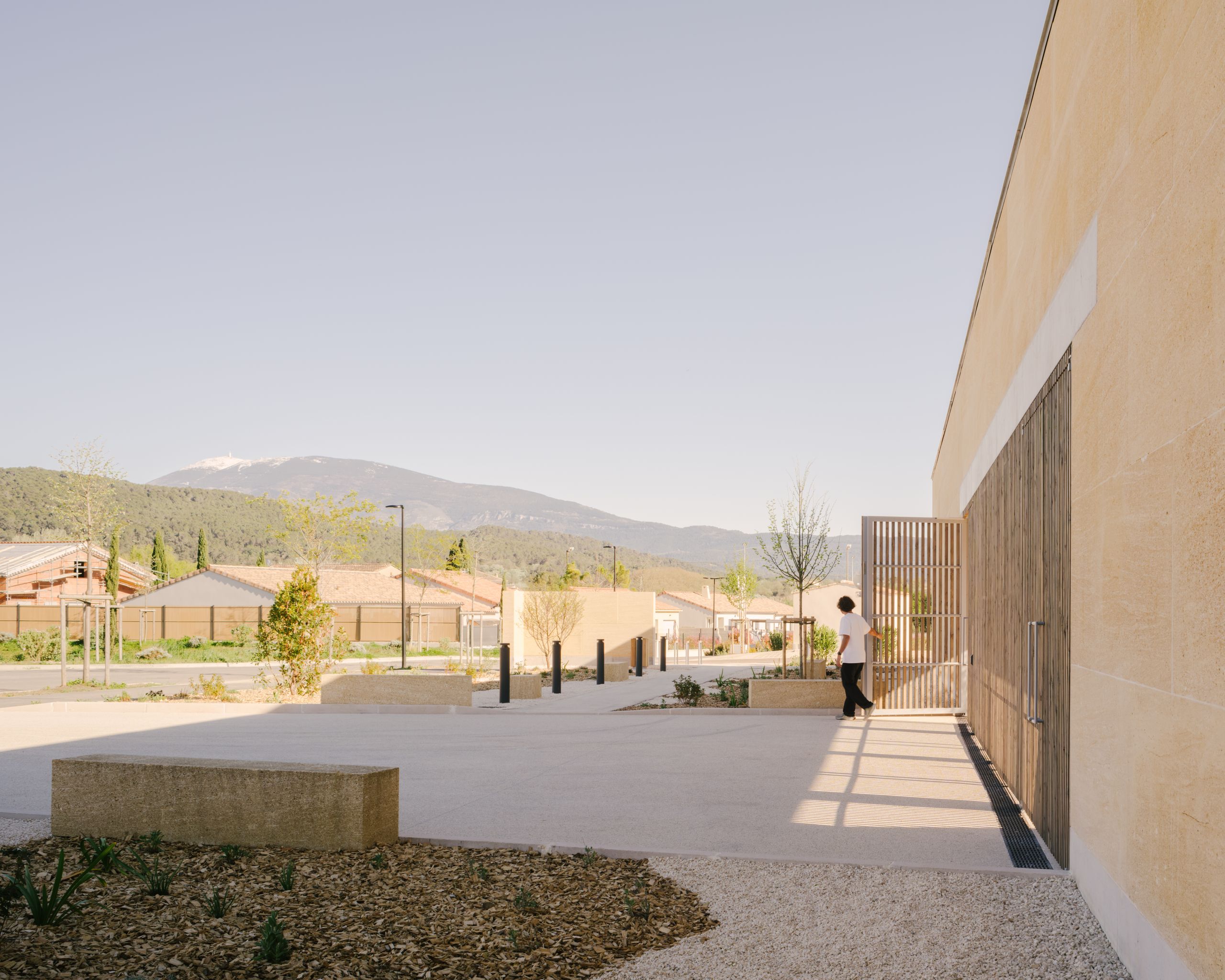 Cour minérale du Pôle culture et jeunesse à Vaison-la-Romaine, cadrée par un mur en pierre massive et des claustras bois, ouverte sur le paysage montagneux.