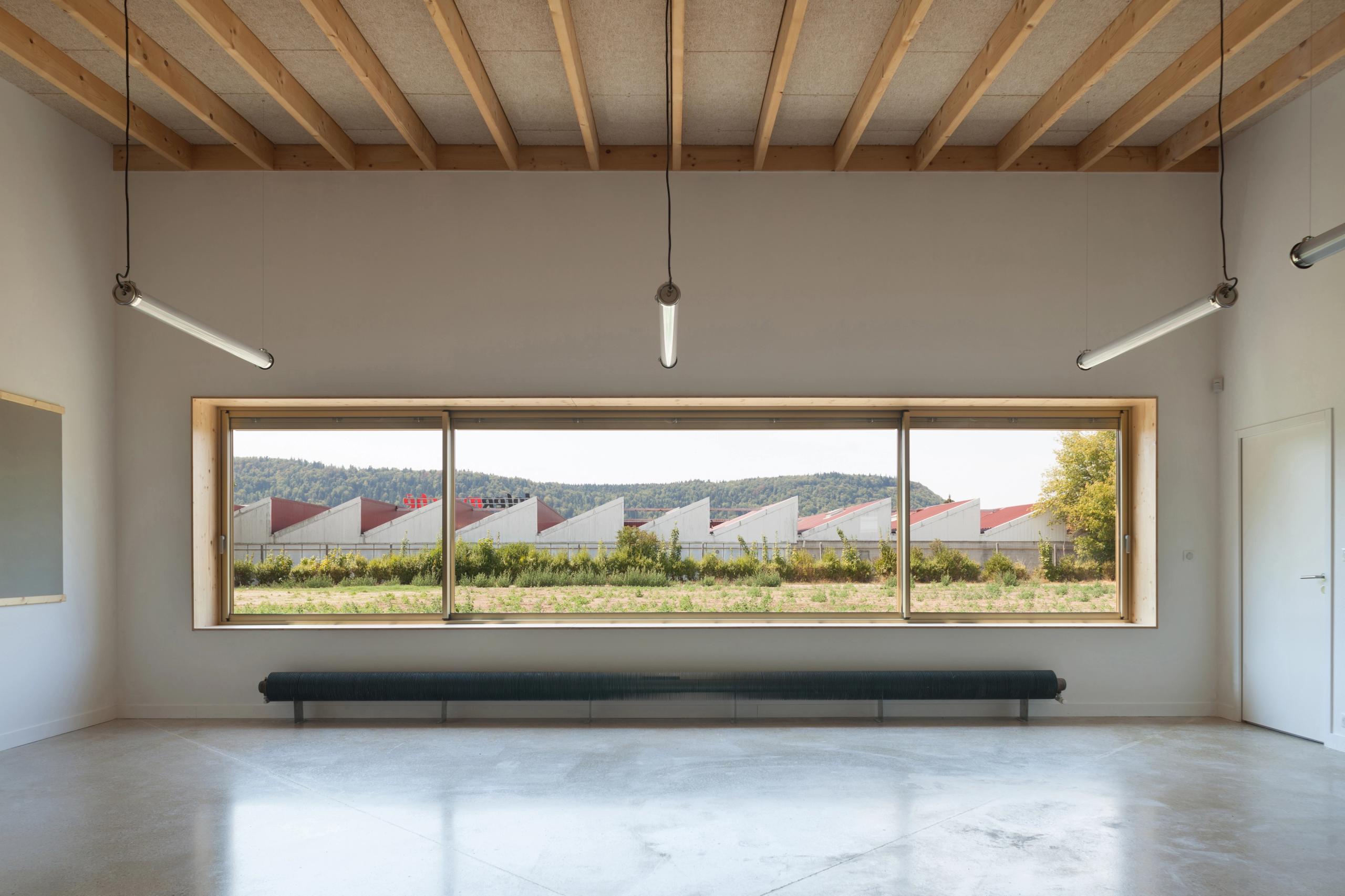 Salle de classe de l’école élémentaire Hubert Reeves à Champagnole, ossature bois apparente, grande baie vitrée cadrant les toitures en sheds et le paysage.