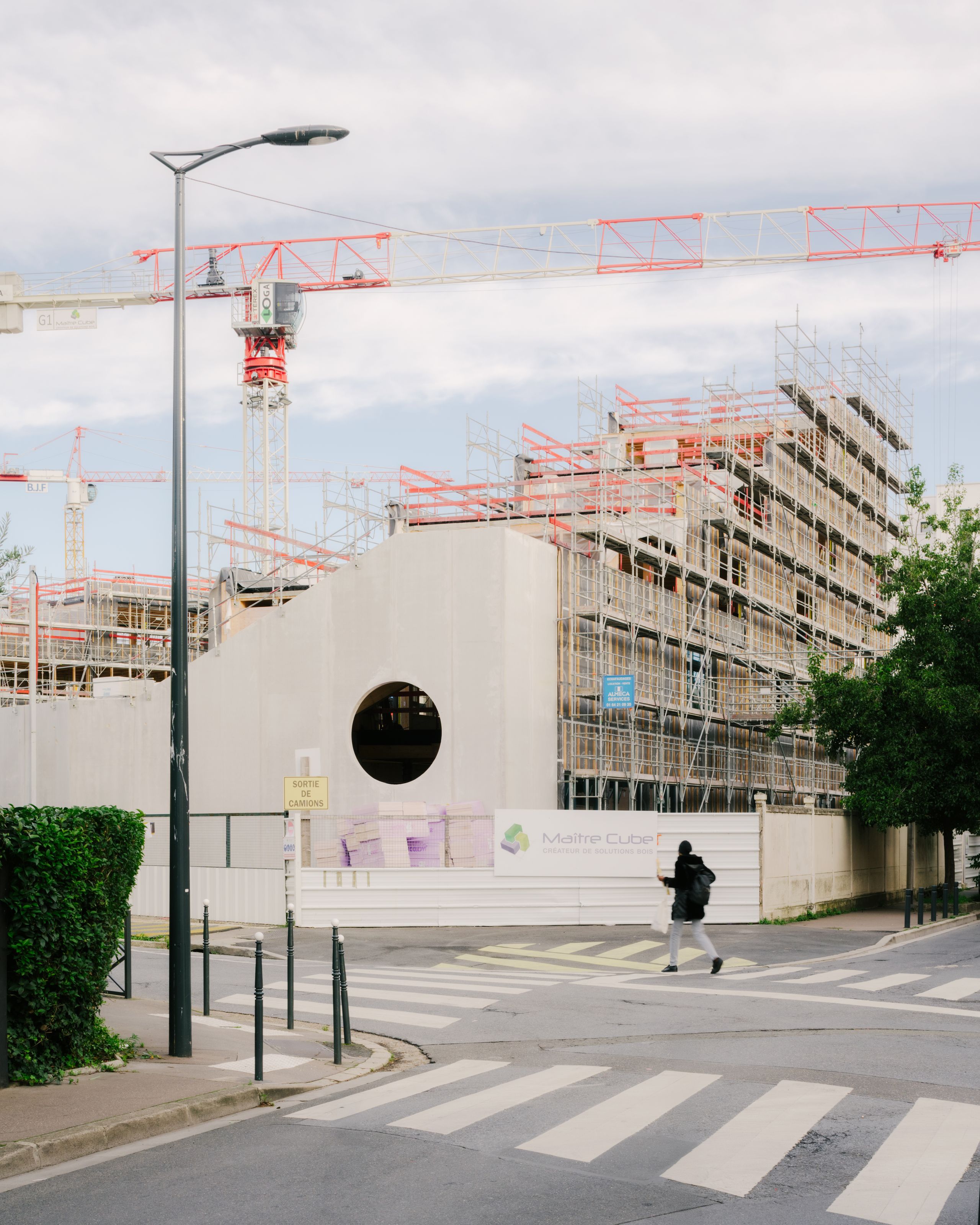 Chantier du groupe scolaire Dominique Frelaut à Colombes : volume en béton percé d’un oculus, façades échafaudées en gradins, grues et tissu urbain dense.