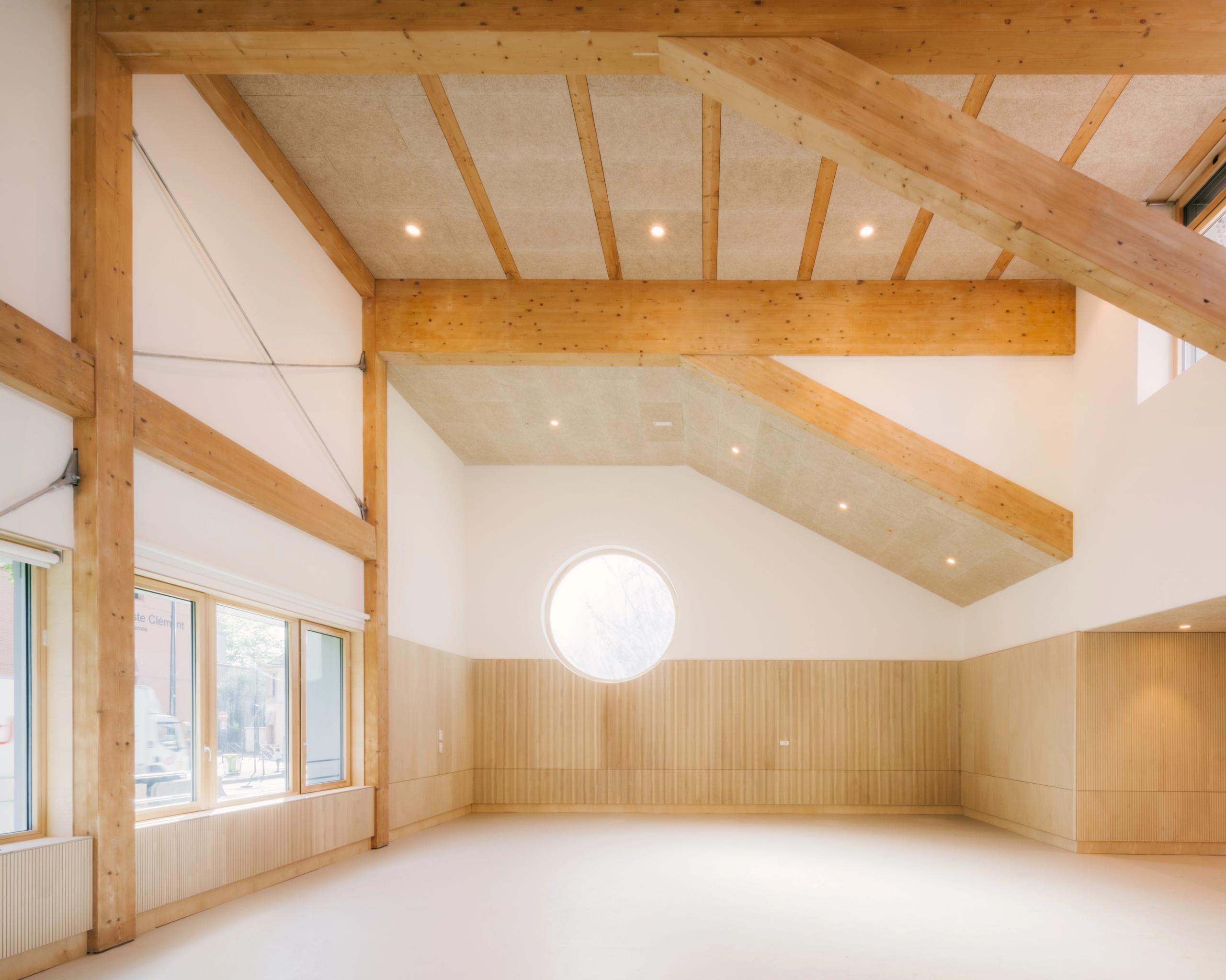 Intérieur du Groupe scolaire Dominique Frelaut à Colombes, grande salle lumineuse en structure bois apparente, plafond incliné et oculus circulaire central.