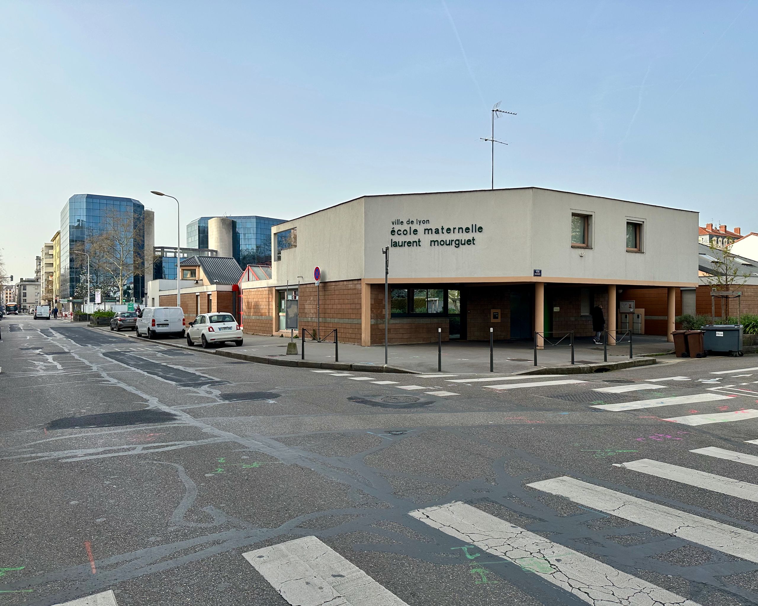 Street view of the Mourguet nursery school in Lyon, a low masonry and rendered volume on a corner plot, forming a sheltered entrance under a recessed ground floor.