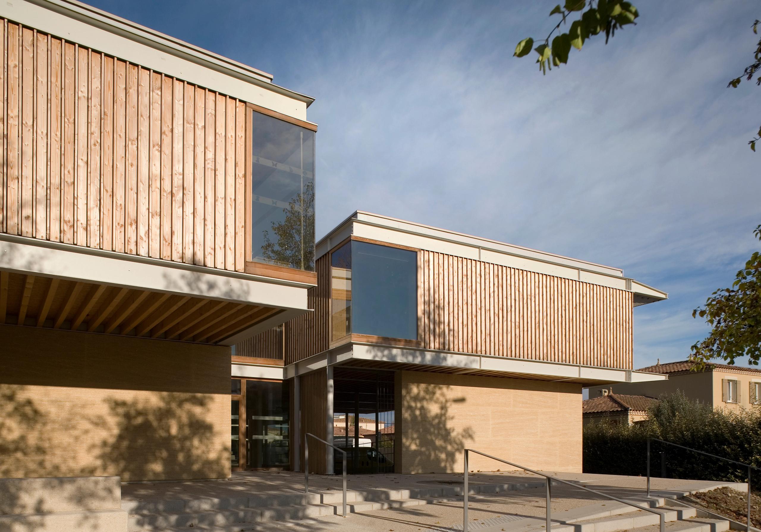 Façade de l’école et centre de loisirs Jean Carrière à Nîmes, volumes sur pilotis en bois et pierre massive, grandes baies vitrées et casquettes métalliques ombragées.