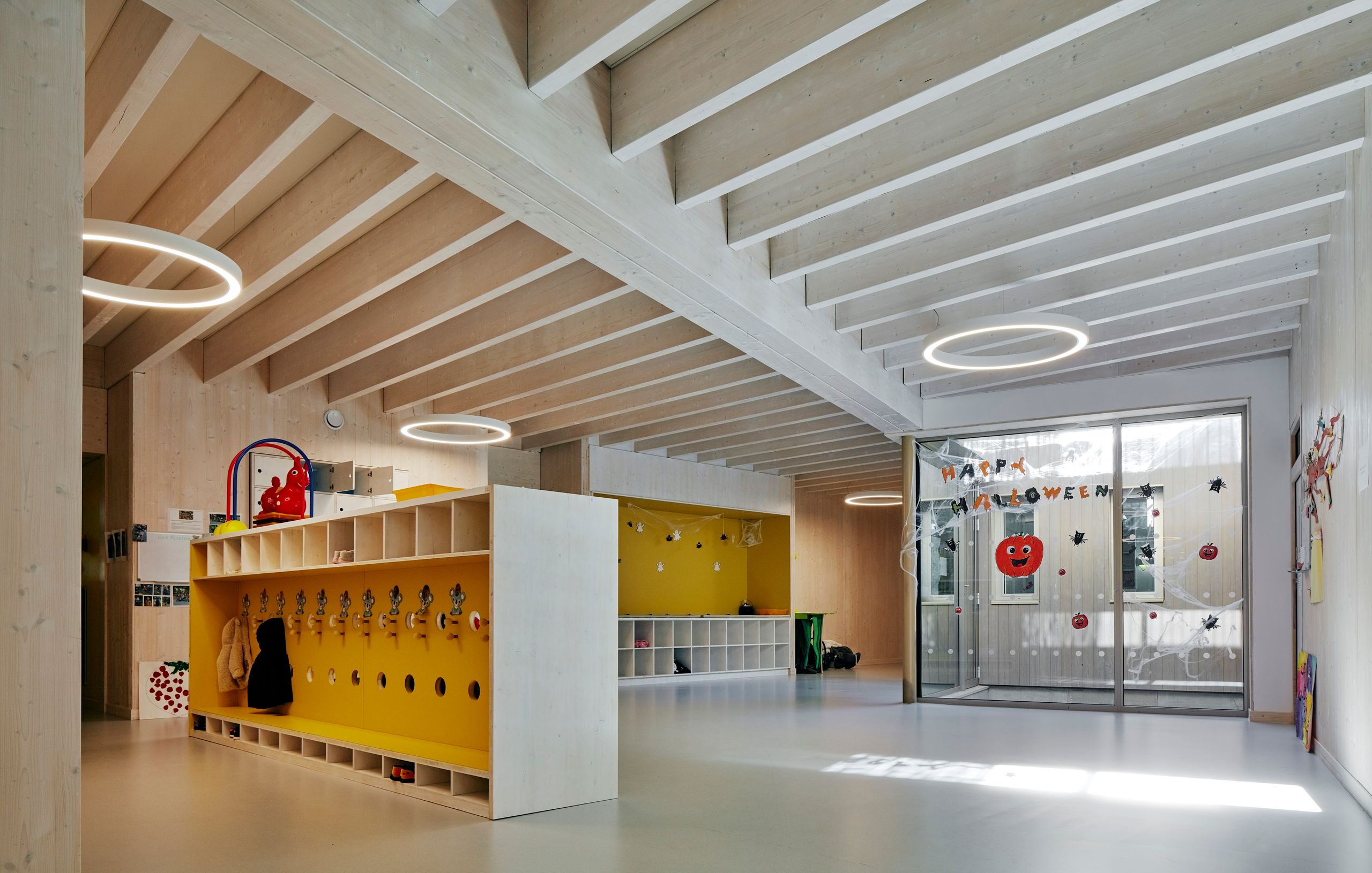Intérieur de la Maison de l’enfance Simone Veil à Albertville : structure bois apparente, volumes ouverts, mobilier bas jaune et grandes baies vitrées vers la cour.